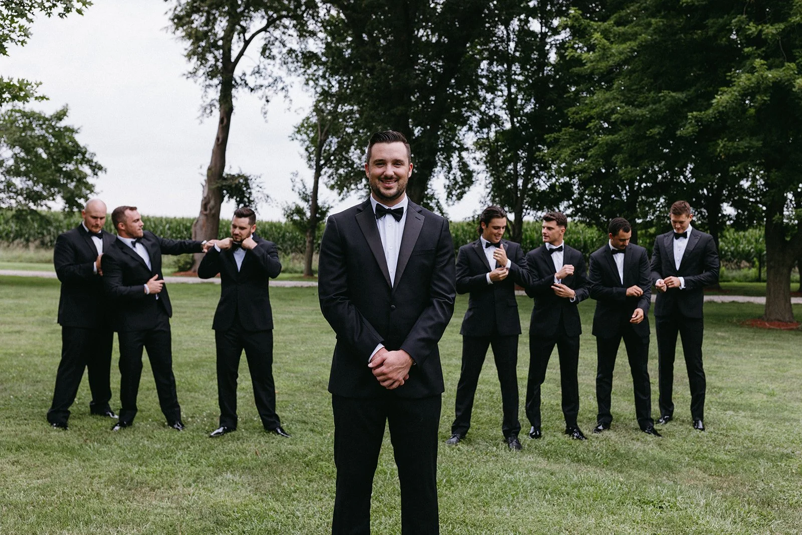 A groom in a black tuxedo stands smiling in front of his groomsmen on a grassy field with trees in the background.