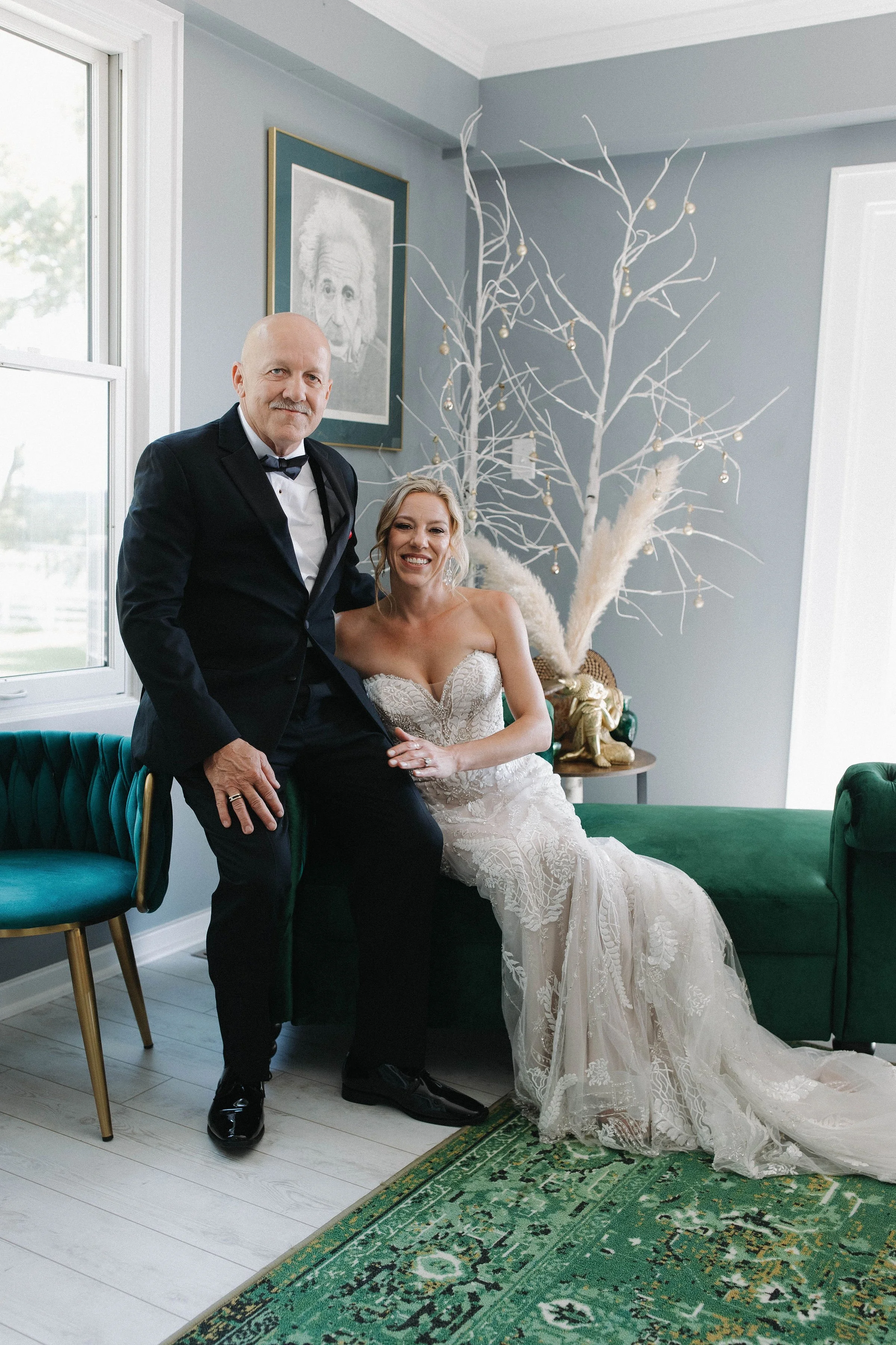 A bride and a groom in formal attire posing together indoors, with a decorative white tree with ornaments behind them, a framed portrait of Albert Einstein on the wall, and a green velvet couch.