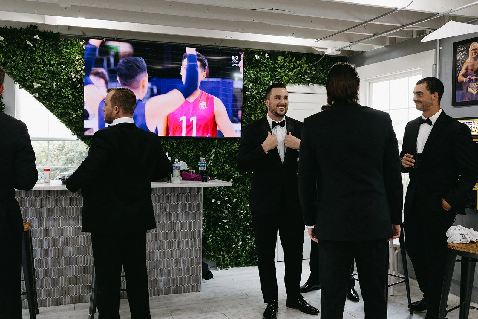 Men in tuxedos socializing at a formal event with a large screen displaying a volleyball game in the background.