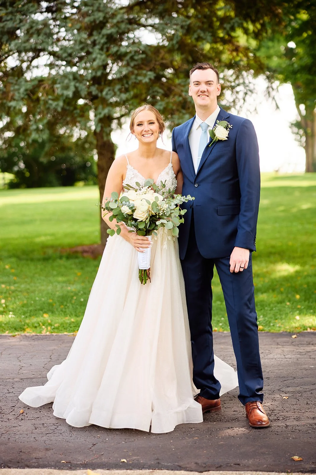 A bride and groom standing outdoors on a wedding day, smiling, with trees and green grass in the background. The bride wears a white wedding dress and holds a bouquet, while the groom wears a blue suit with a white shirt, tie, and boutonniere.