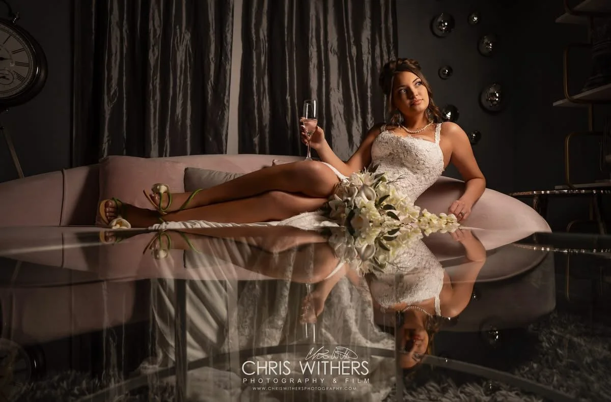 A woman in a wedding dress reclining on a sofa, holding a glass of champagne, with a bouquet of flowers in her lap, in a modern, dimly lit room.