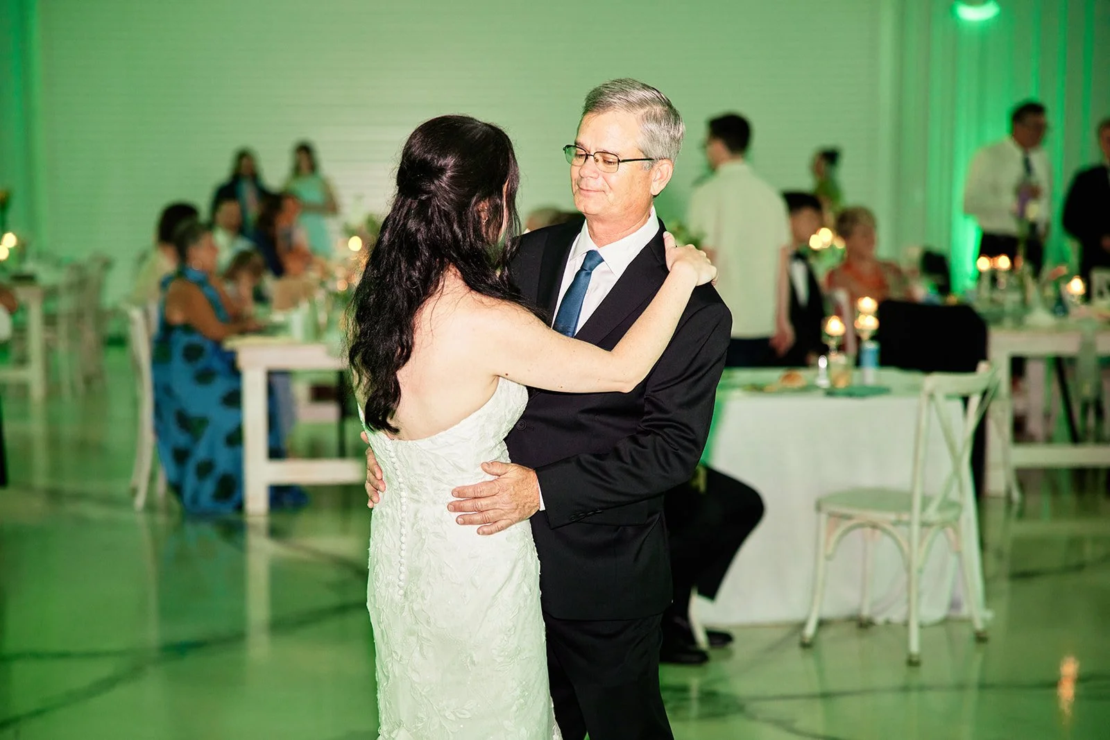 A bride and groom dancing at a wedding reception in a decorated venue with tables and guests in the background.