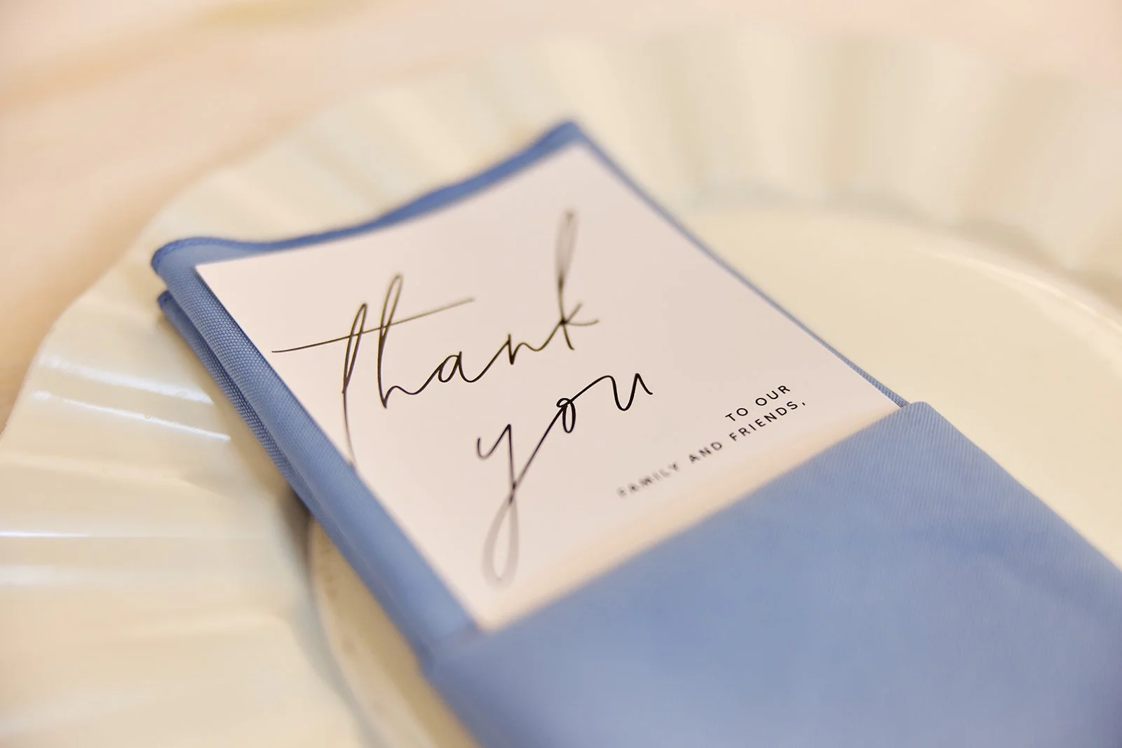 A thank you card with elegant black cursive text that reads 'Thank you' on a white background, with smaller text below that says 'to our family and friends.' The card is surrounded by a blue fabric sleeve or holder, resting on a white, textured surfa