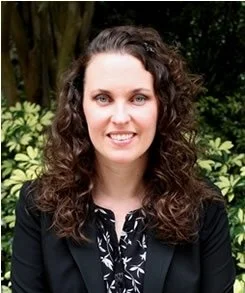 Person with curly brown hair, wearing a black blazer and patterned blouse, standing in front of leafy green plants.