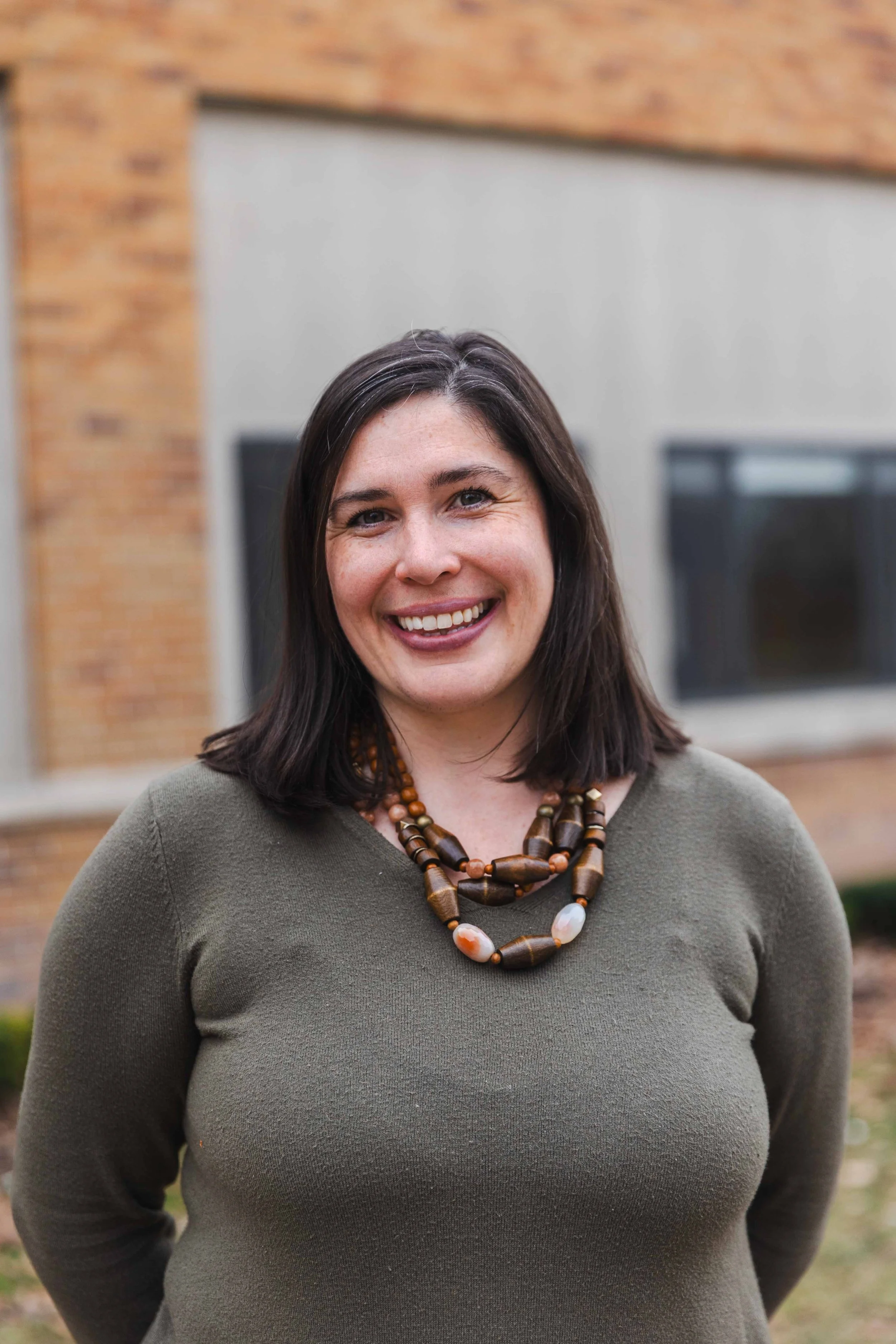 Smiling person wearing a green sweater and a beaded necklace standing outside a brick building.