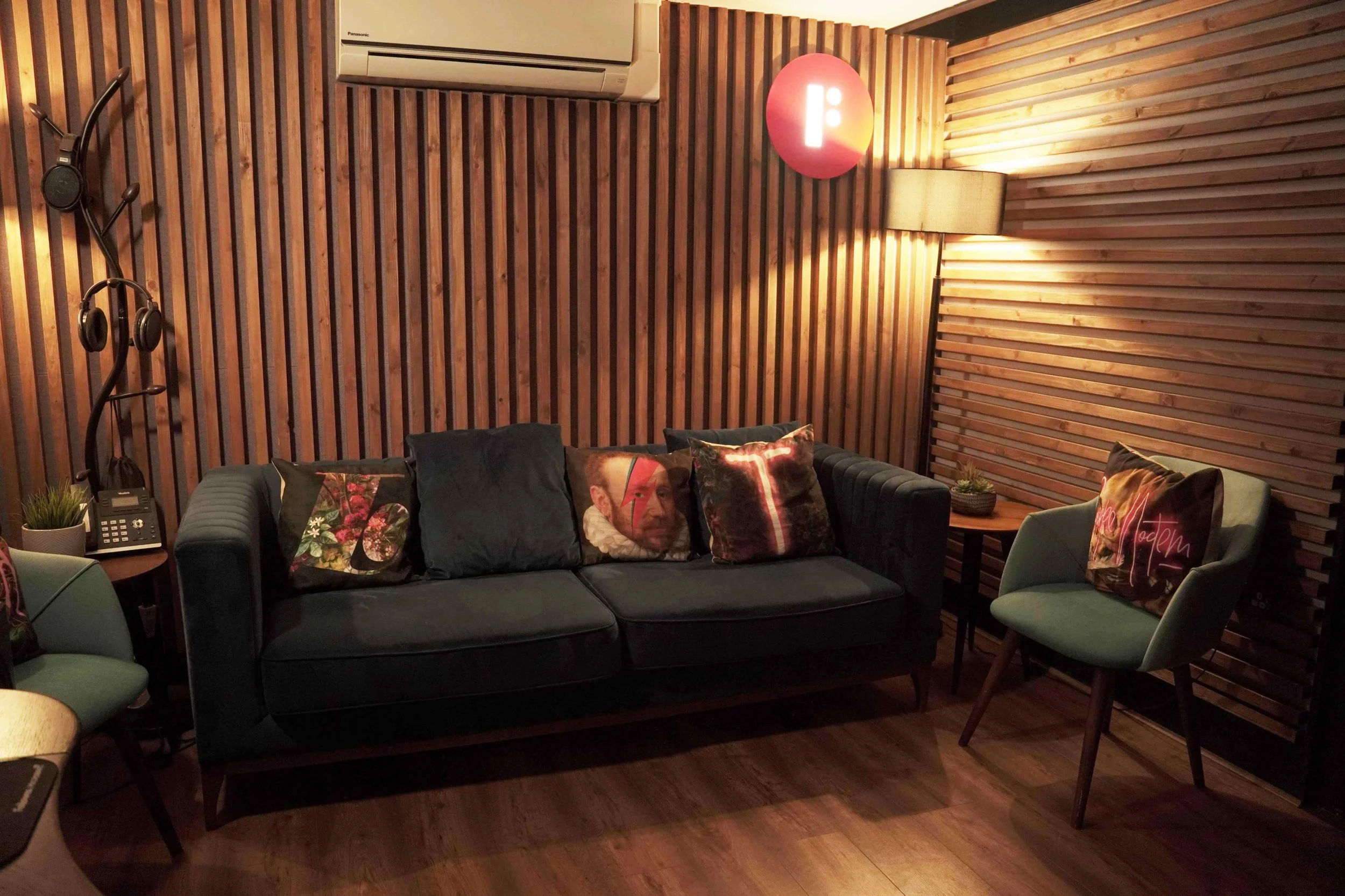 A cozy living room with wooden paneled walls and warm lighting. There are a black sofa with decorative pillows, a mint green chair, a small side table with a plant, a lamp, and a coat rack. An air conditioning unit is mounted on the wall, and a pink ceiling light provides additional lighting.