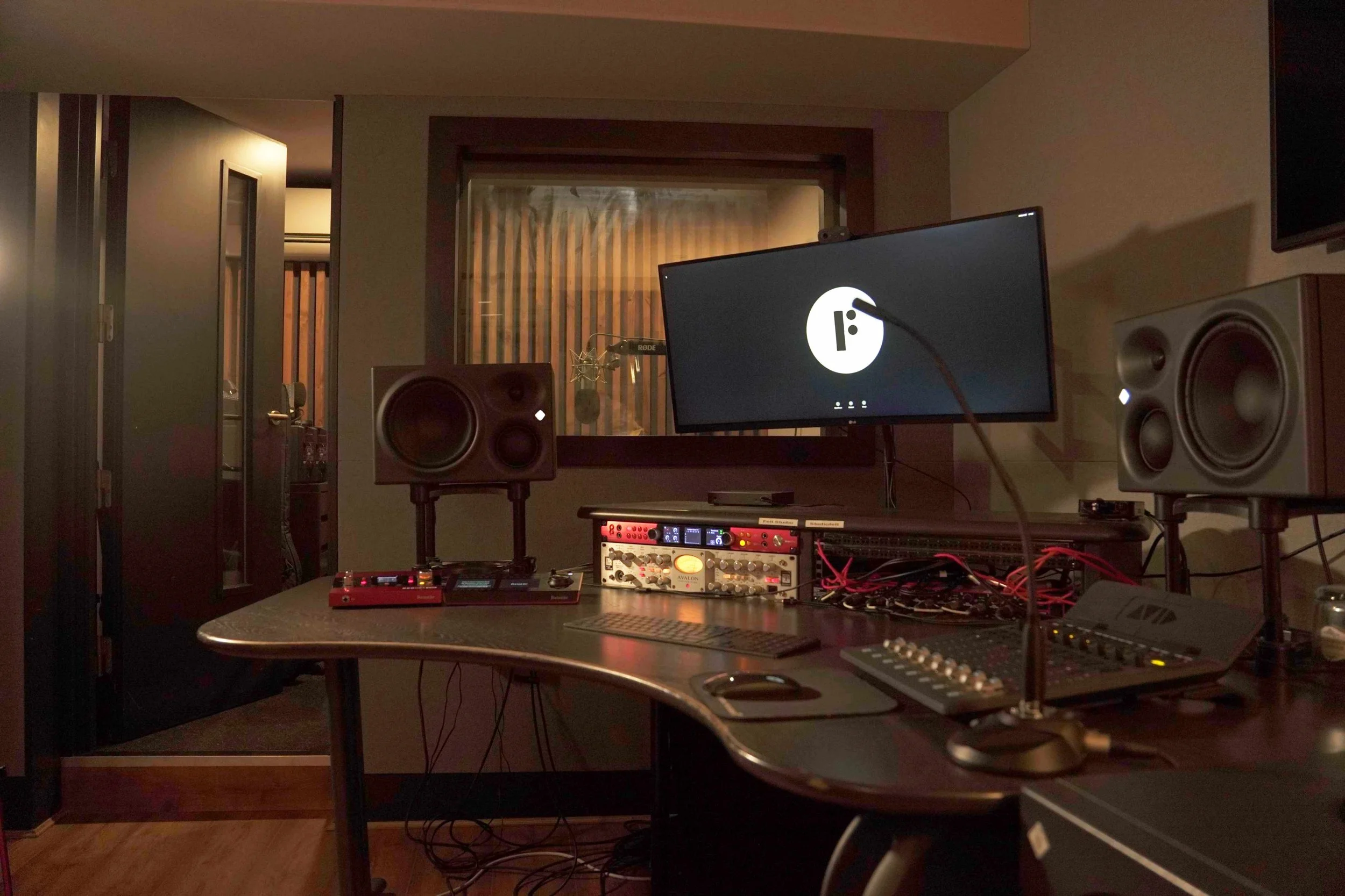 Recording studio setup with computer monitors, studio speakers, audio interface, and musical equipment on a desk in a dimly lit room.