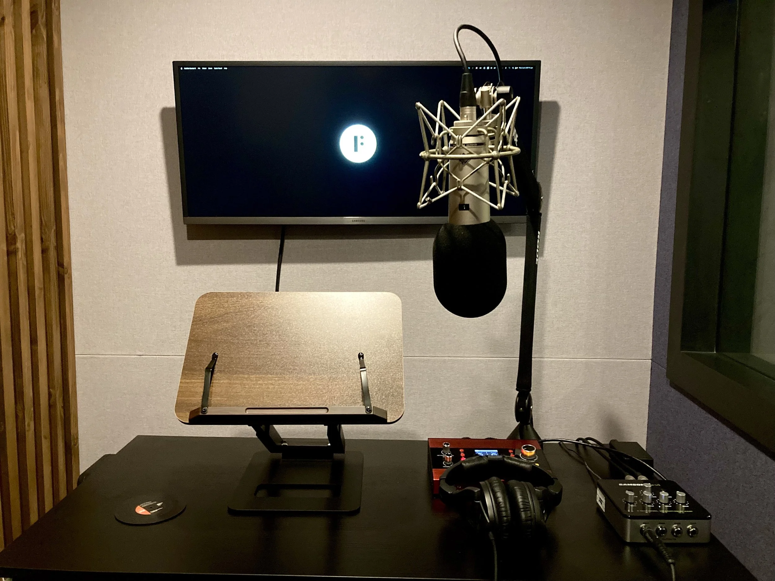 Music recording studio setup with a television, microphone in a shock mount, headphones, audio mixer, and a imported MIDI controller on a dark table.