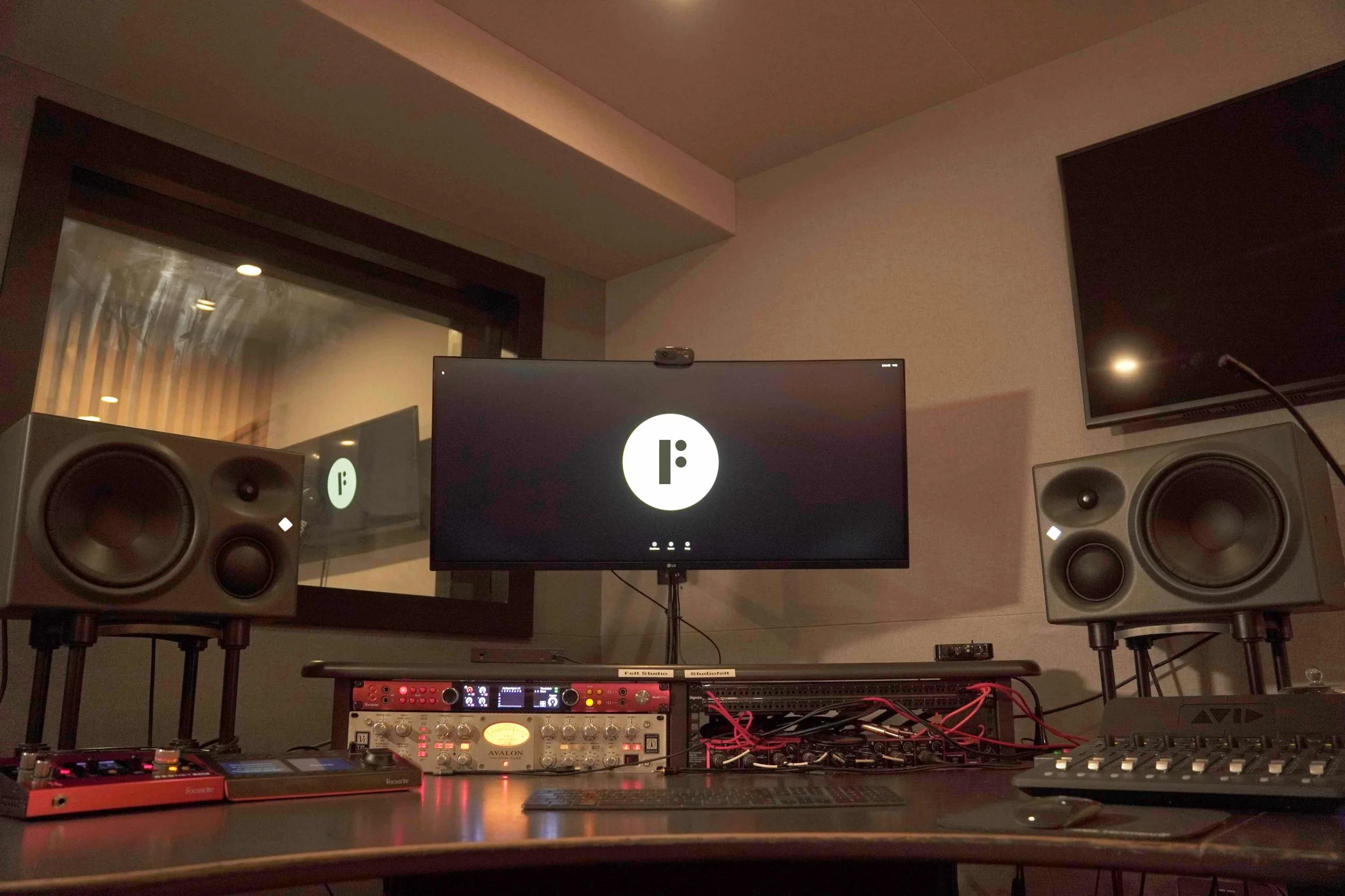 Recording studio with large studio monitors, audio equipment, a computer monitor displaying a logo, and a mixing console on a desk.