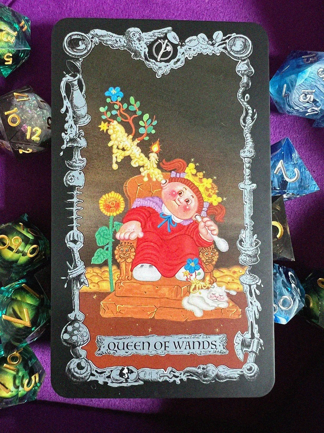 A Queen of Wands card from the Garbage Pail Kids deck sits on a bed of different polyhedral dice. She sits on a throne with lion motifs, with an earwax covered q-tip going through her head via the ears, and a cat with a similar q-tip at her feet.