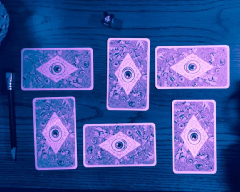 A selection of six tarot cards laid out on a wooden desk with a pencil and d6 die. The photo is grainy and is edited to have shades of purples and blues.