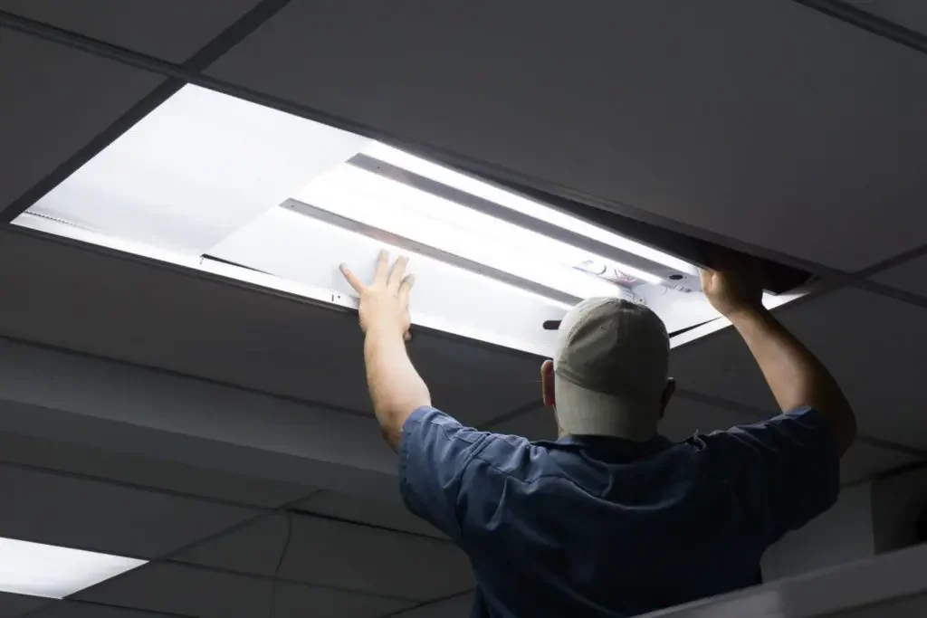 A person in a baseball cap and dark shirt is installing or replacing a fluorescent light fixture in a ceiling with drop ceiling tiles.