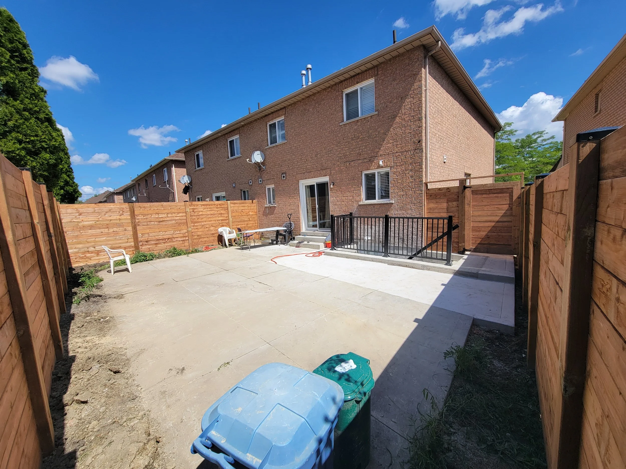 Backyard with concrete patio, wooden fence, and two-story brick townhouse. Plastic bin and chair visible; clear blue sky.