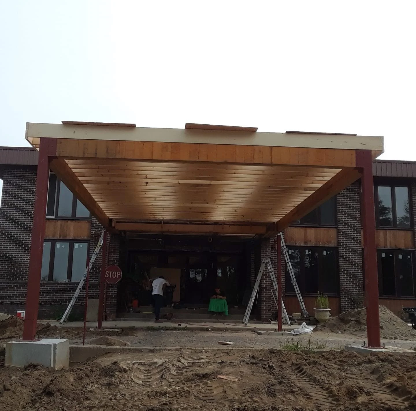 Construction of a porch or entrance canopy outside a brick building, featuring wooden beams and metal support columns, with construction materials and ladders visible.