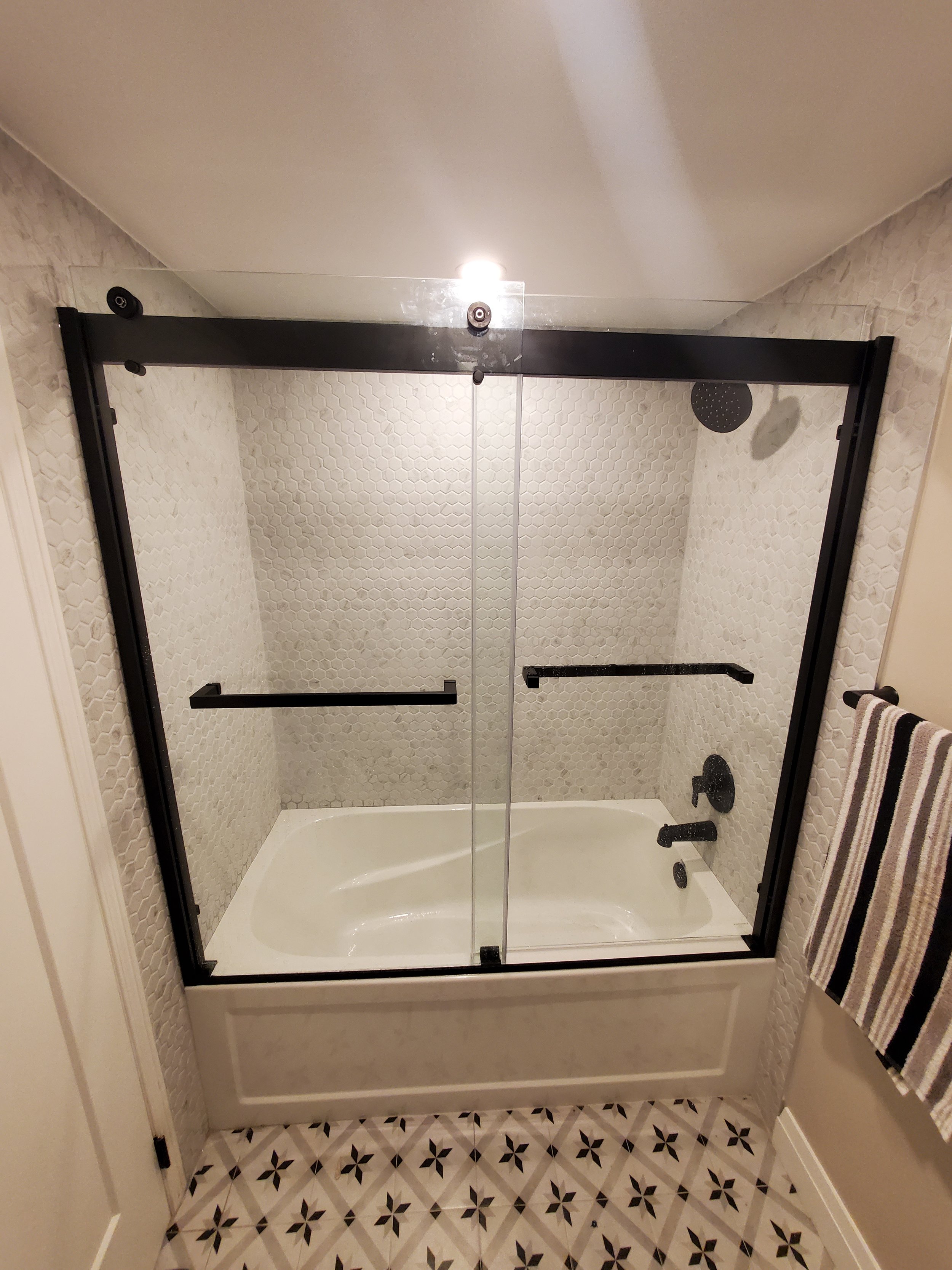 Modern bathroom featuring a white bathtub enclosed with a glass sliding door, black fixtures, geometric pattern floor tiles, and a striped towel on a bar.