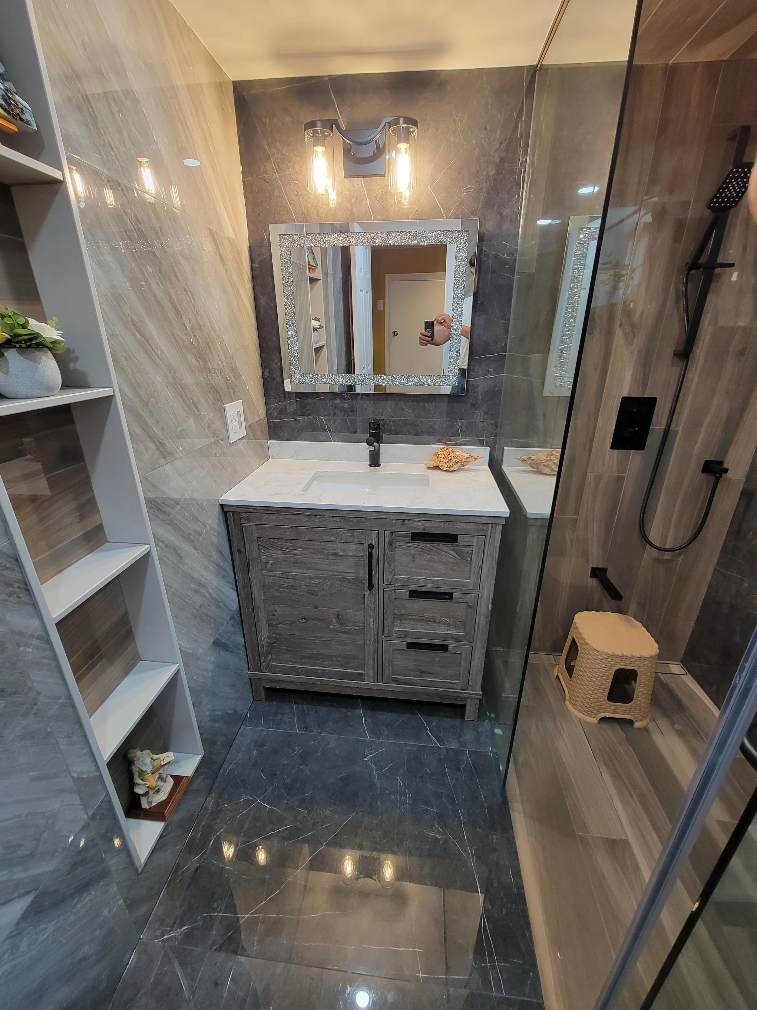Modern bathroom with gray marble floors and walls, a wooden vanity with a white countertop, a decorative mirror, and a glass shower enclosure. The space includes shelving with decor and wall-mounted lighting fixtures.