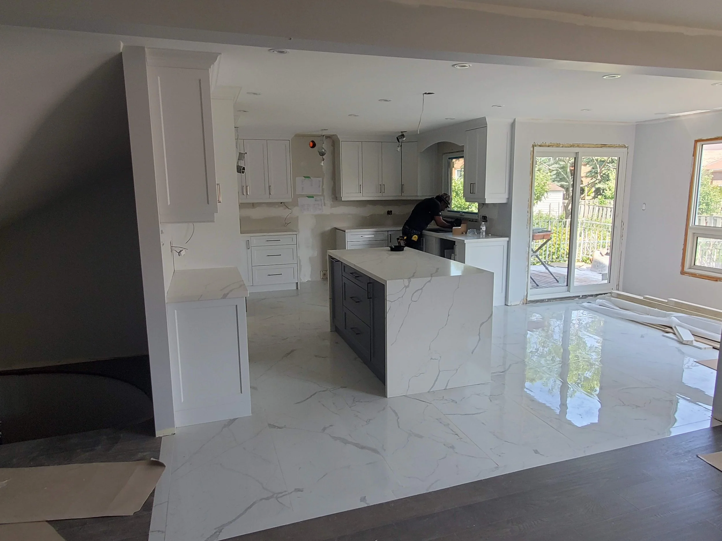 Modern kitchen under renovation with white cabinets, marble countertops, and center island. Person working near sliding glass doors.