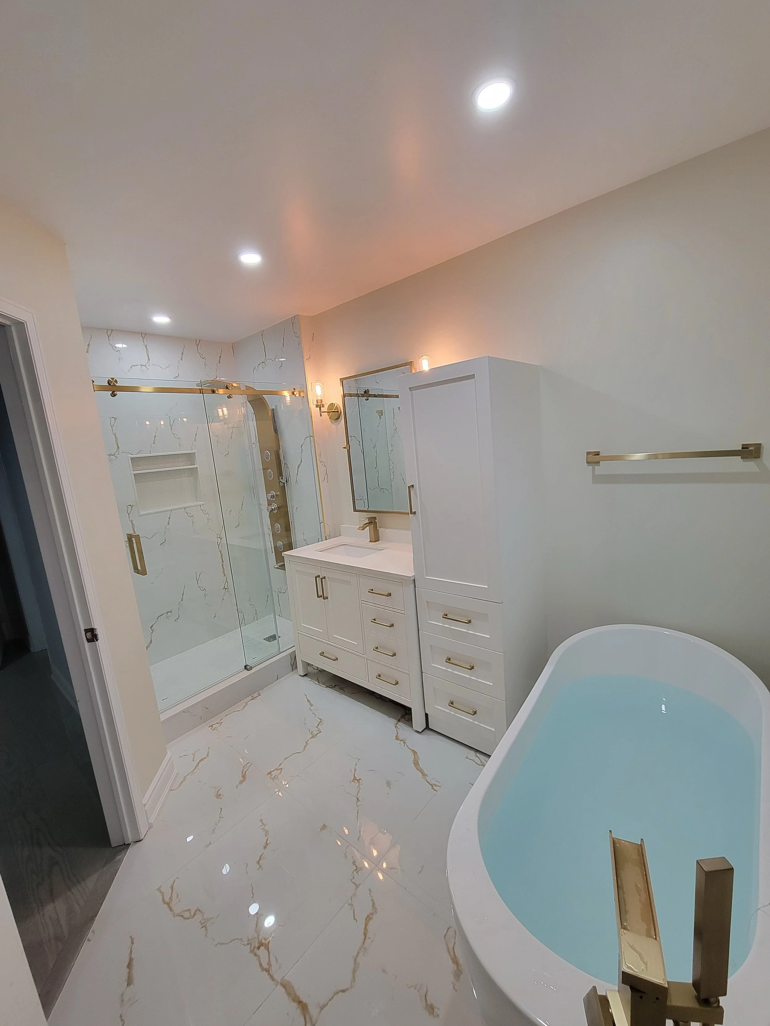 Modern bathroom with marble tiles, a glass-enclosed shower, freestanding bathtub, white vanity with gold fixtures, and recessed lighting.