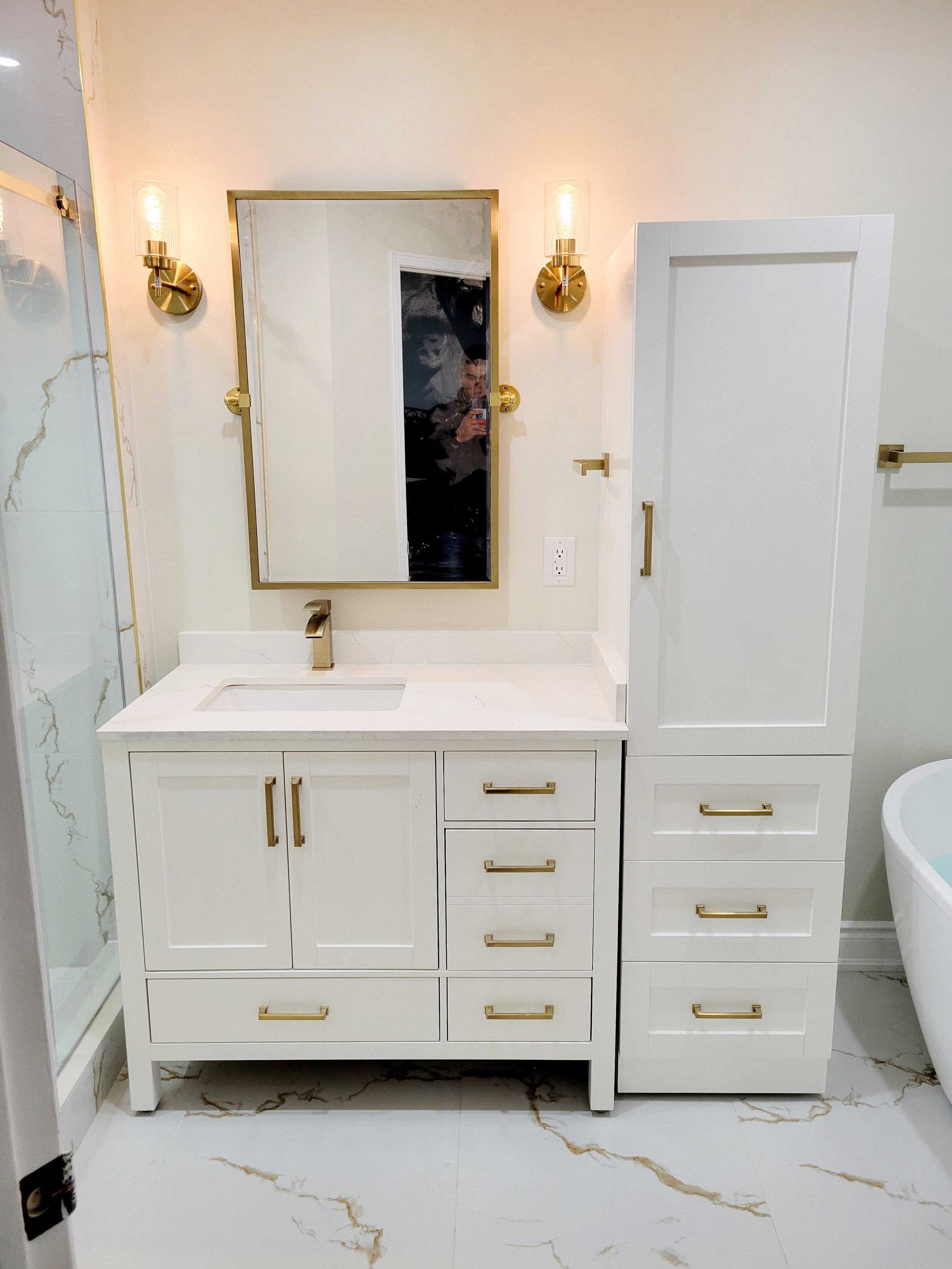 Modern bathroom vanity with white cabinets, gold hardware, a rectangular mirror, and two wall sconces. The floor and shower feature marble-like patterns. A portion of a bathtub is visible on the right.