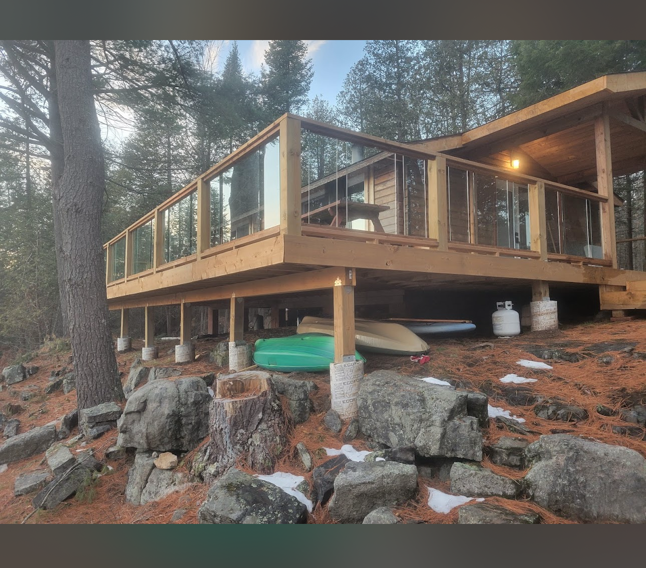 A wooden cabin with a raised deck and glass railings in a forest setting, surrounded by trees and rocky terrain. Canoes and a propane tank are stored underneath the deck.