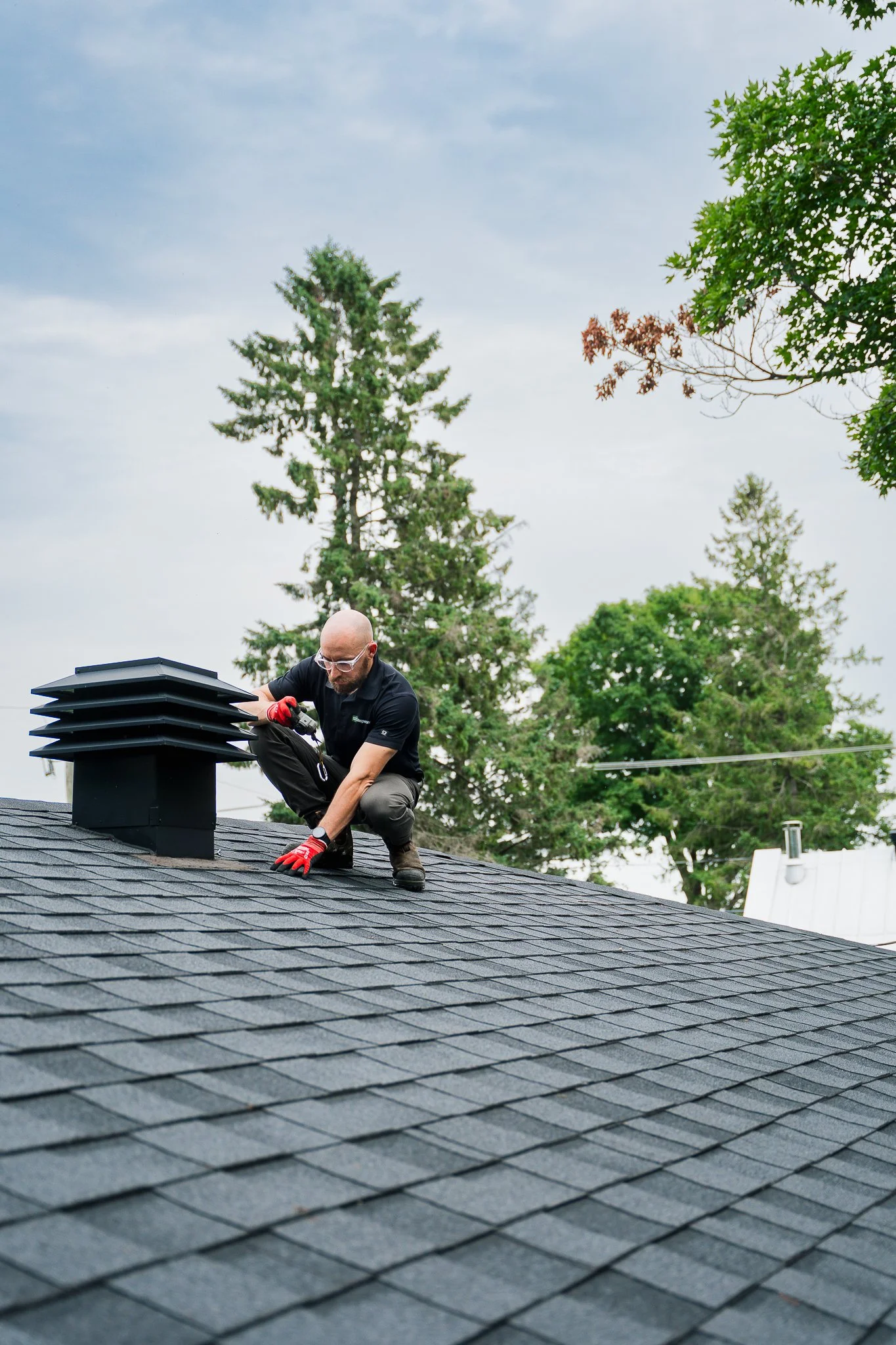Un homme portant des lunettes de protection, des gants rouges, travaille sur le toit d'une maison, posant ou inspectant des tuiles de toiture noires. Il y a des arbres en arrière-plan et un ciel nuageux.