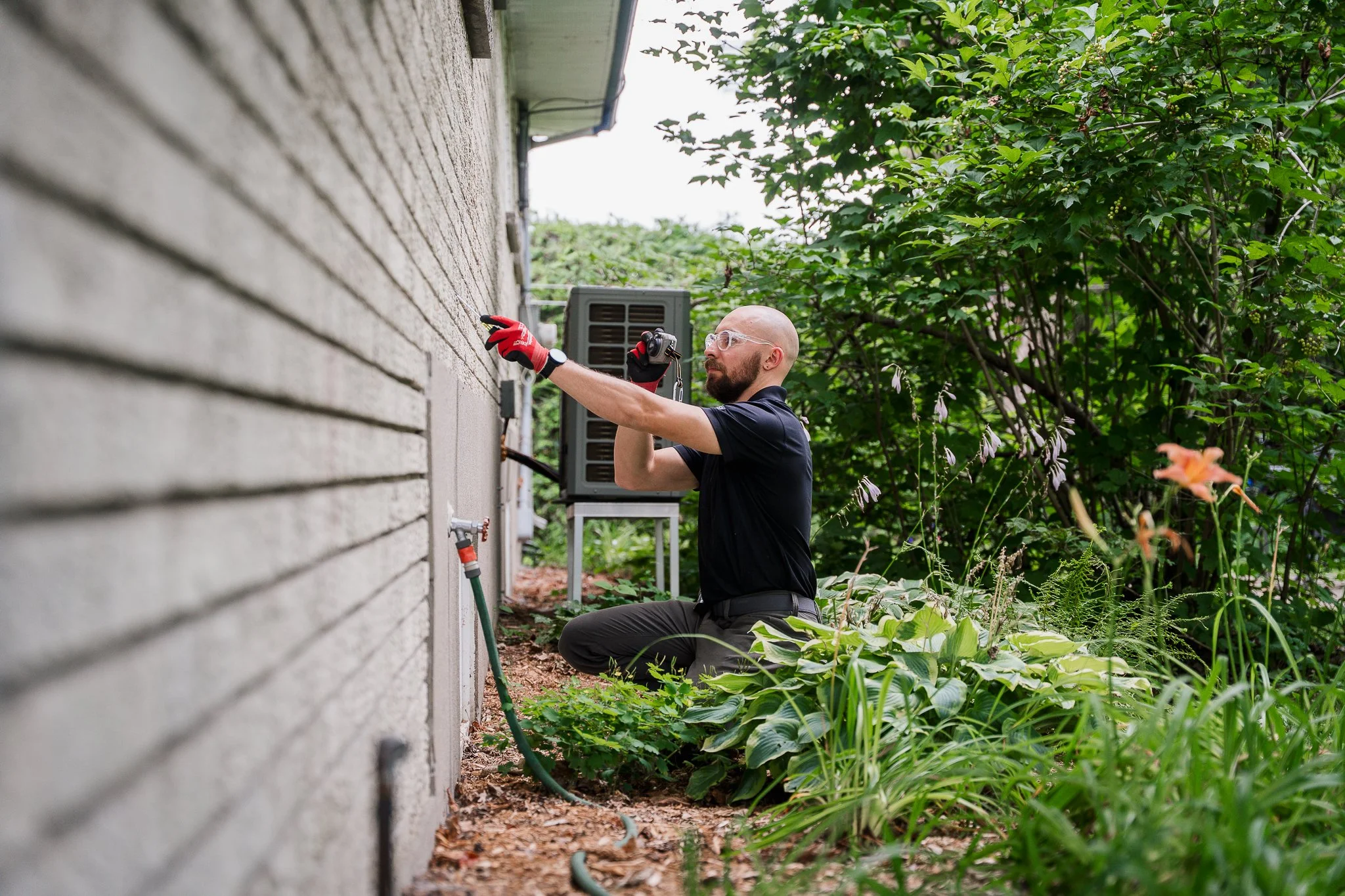 Un homme qui s'agenouille à côté d'une maison, inspectant des câbles ou équipements électriques fixés au mur, entouré de verdure, portant des lunettes de sécurité, une montre et des gants rouges.