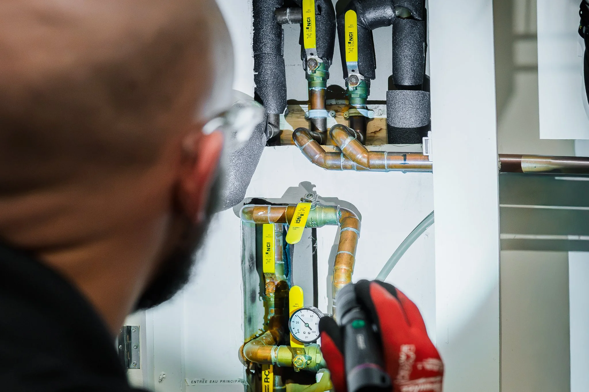 Technicien utilisant un outil électrique pour travailler sur un système de plomberie comprenant des tuyaux en cuivre et des vannes avec étiquettes jaunes.