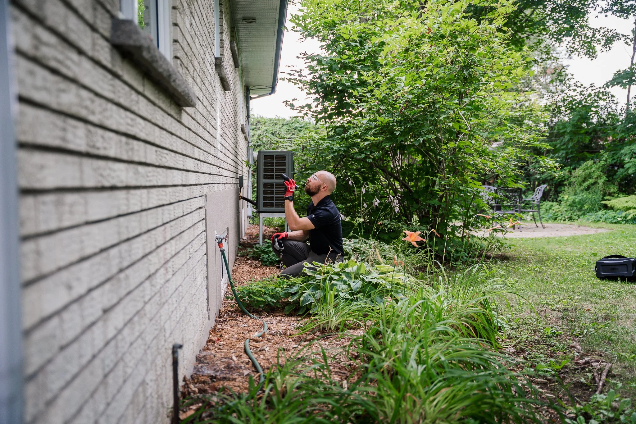 Un homme en T-shirt noir et lunettes, portant des gants rouges, examine ou travaille à côté du mur extérieur d'une maison, dans un jardin avec des arbustes et des plantes vertes, utilisant un outil électrique