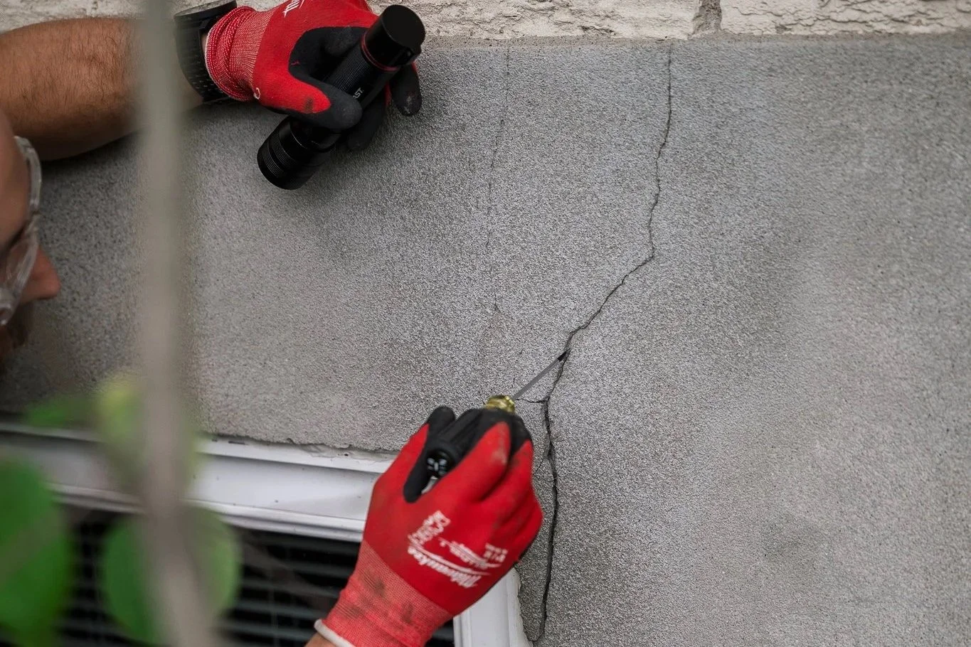 Une personne portant des gants rouges effectue une réparation de fissure sur un mur en béton en utilisant un outil de scellement ou de mastic.