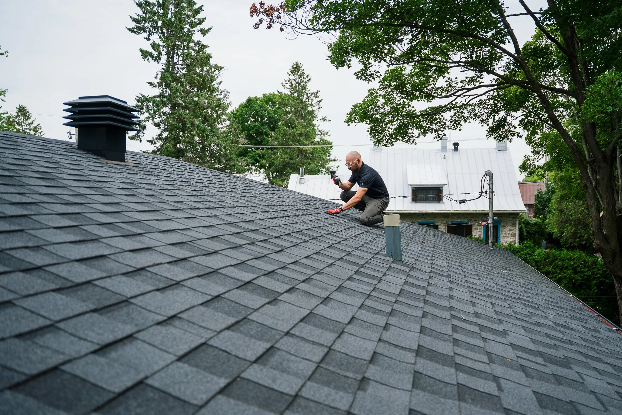 Un homme en vêtements noirs et lunettes travaille sur le toit en ardoises d'une maison, entouré d'arbres et de maisons voisines, sous un ciel gris.