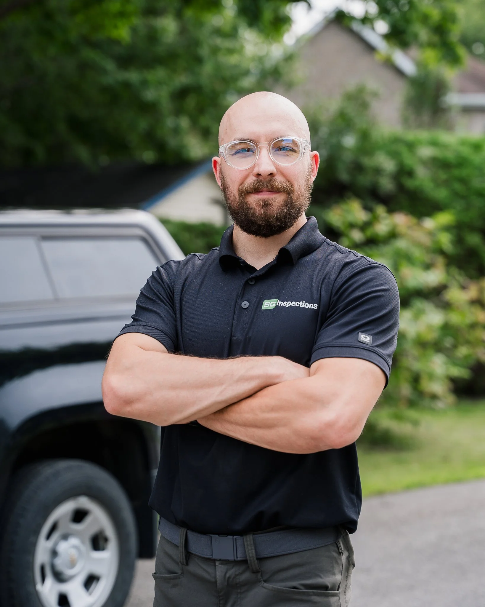 Homme portant des lunettes transparentes et un polo noir avec un logo 'SG Inspections', bras croisés, debout devant une voiture et un paysage verdoyant.
