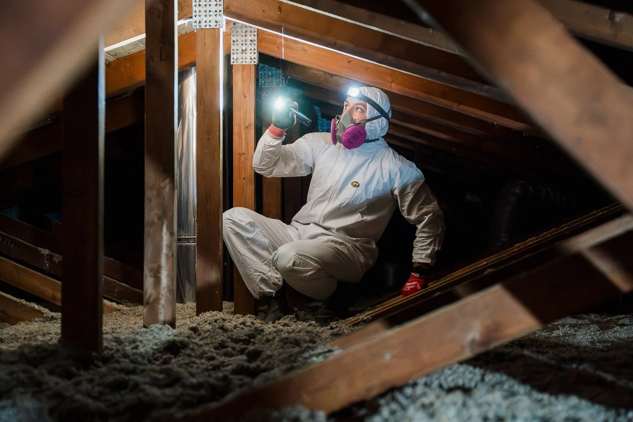 Une personne habillée en combinaison de protection, portant un masque et une lampe frontale, inspecte une zone sous un toit en bois dans une grenier, probablement pour des travaux de rénovation ou d'isolation.
