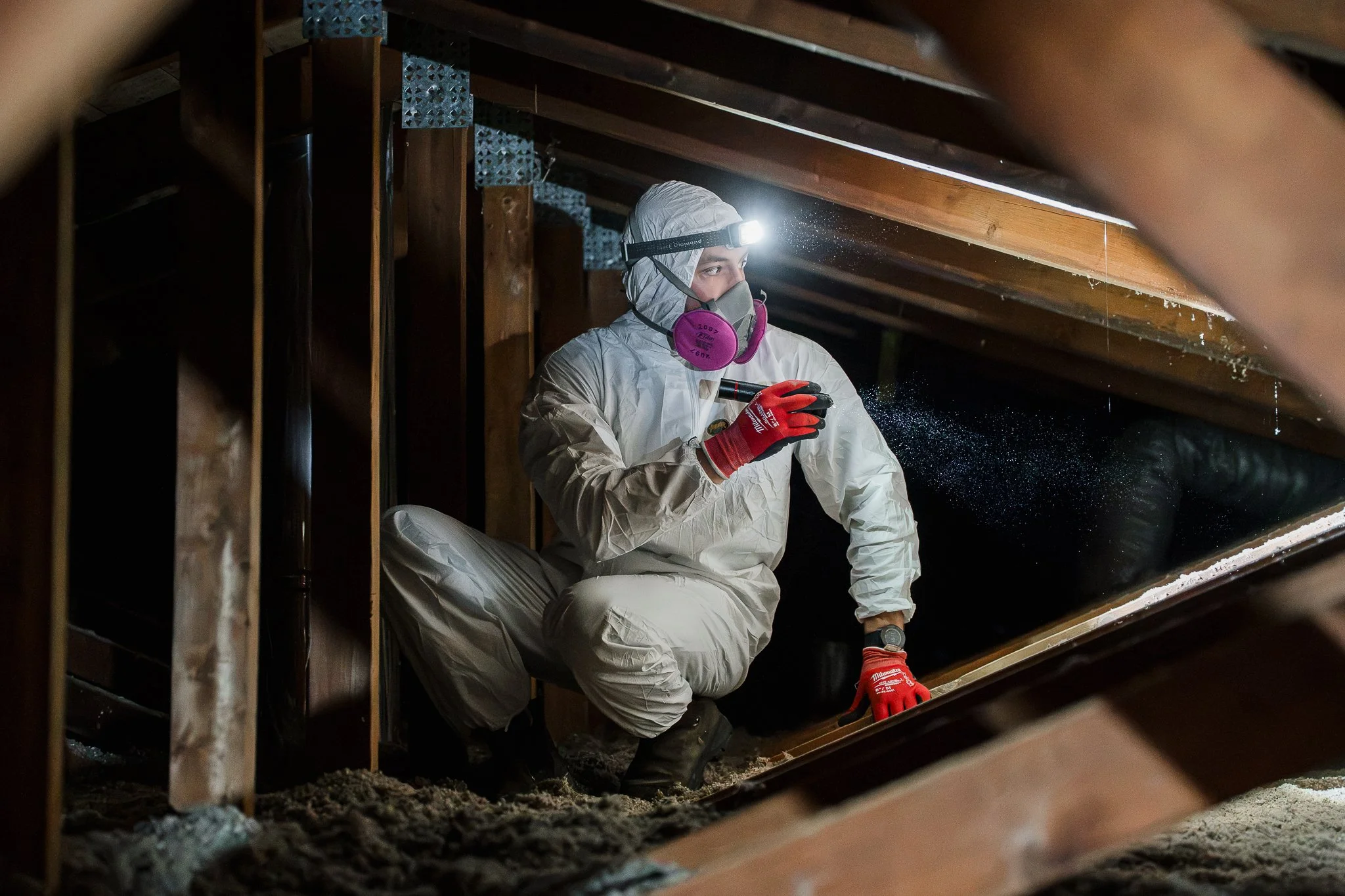 Une personne portant des vêtements de protection, une lampe frontale et un masque, examine une zone sous un toit en bois, probablement en train d'effectuer des travaux ou une inspection dans un environnement sombre.