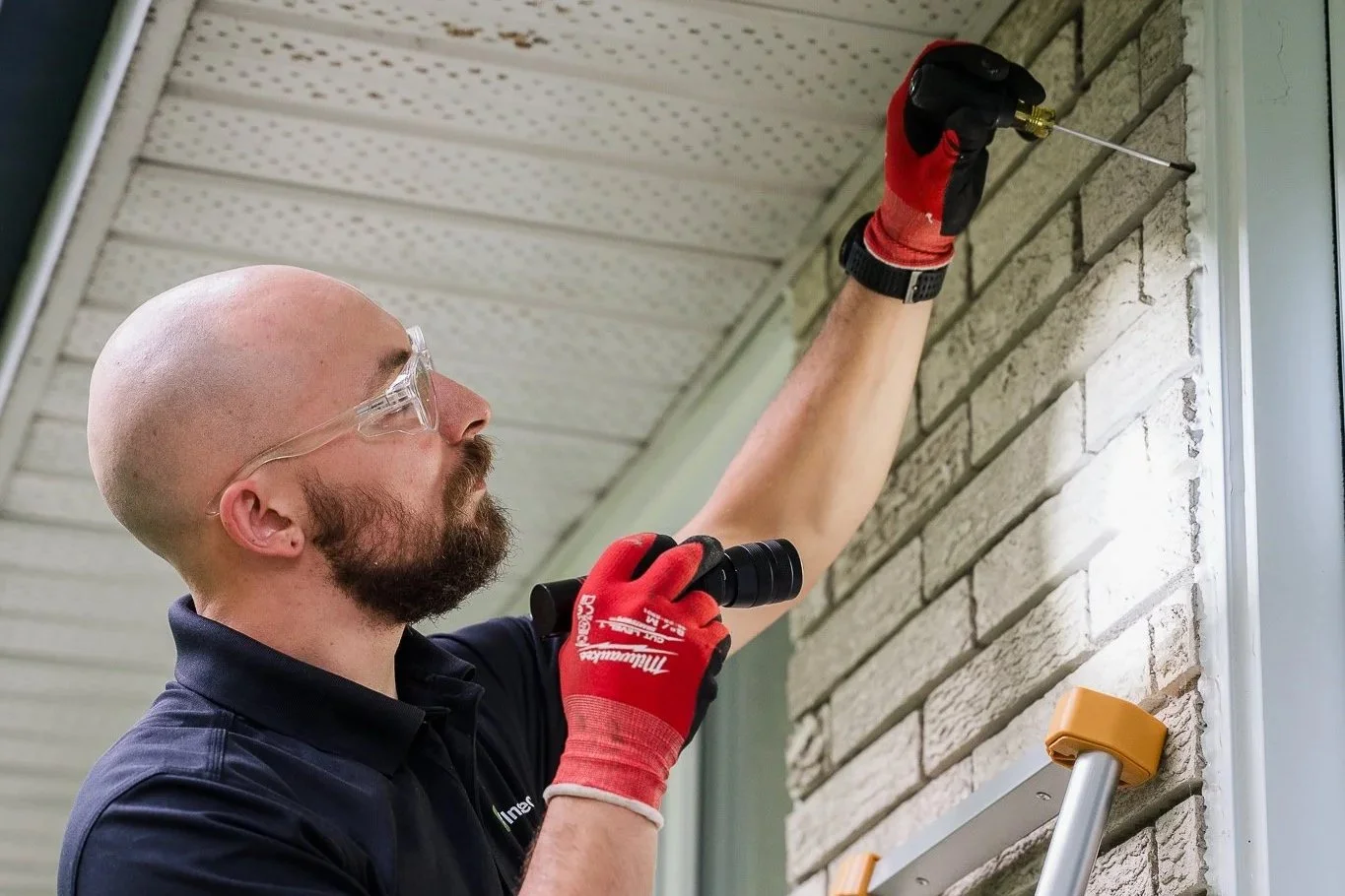 Un homme portant des lunettes de protection et des gants rouges utilise un tournevis pour fixer une pièce au mur en briques gris clair, avec une échelle en dessous.