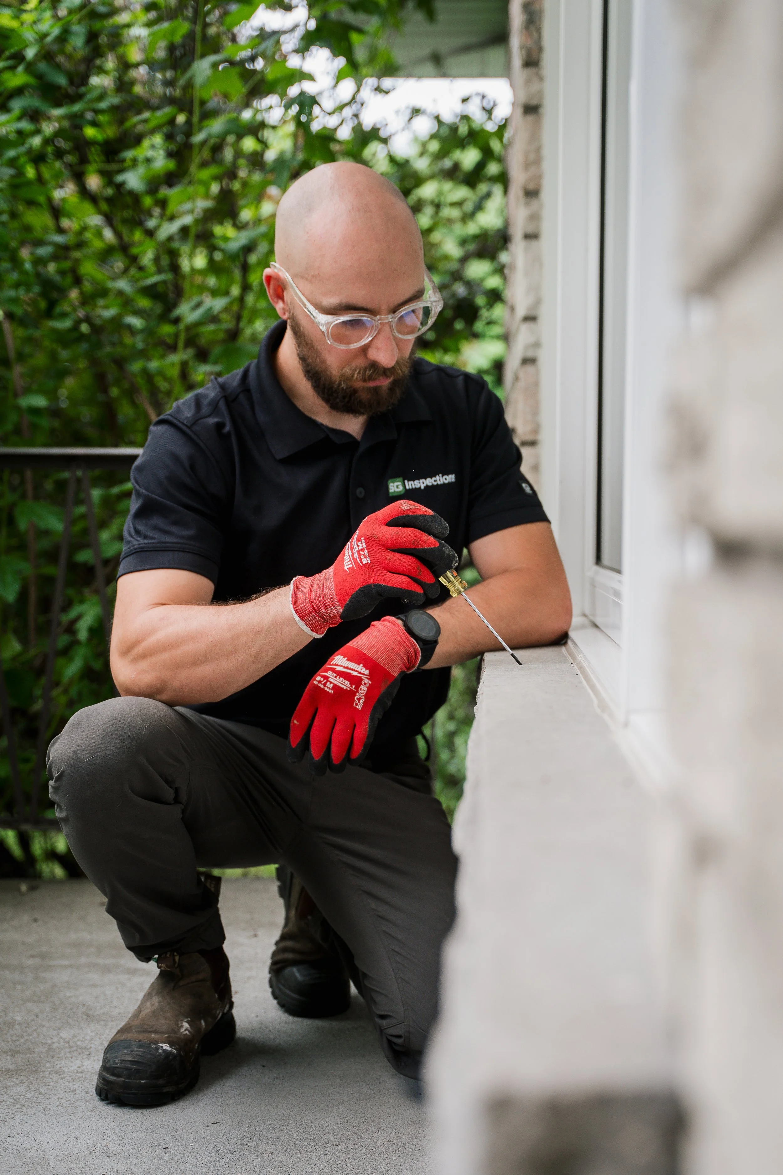 Un homme casqué avec des lunettes transparentes, portant un polo noir avec un logo, est à genoux en train d'inspecter ou de réparer une boîte aux lettres extérieure en utilisant un tournevis. Il porte des gants rouges et des bottes de travail.