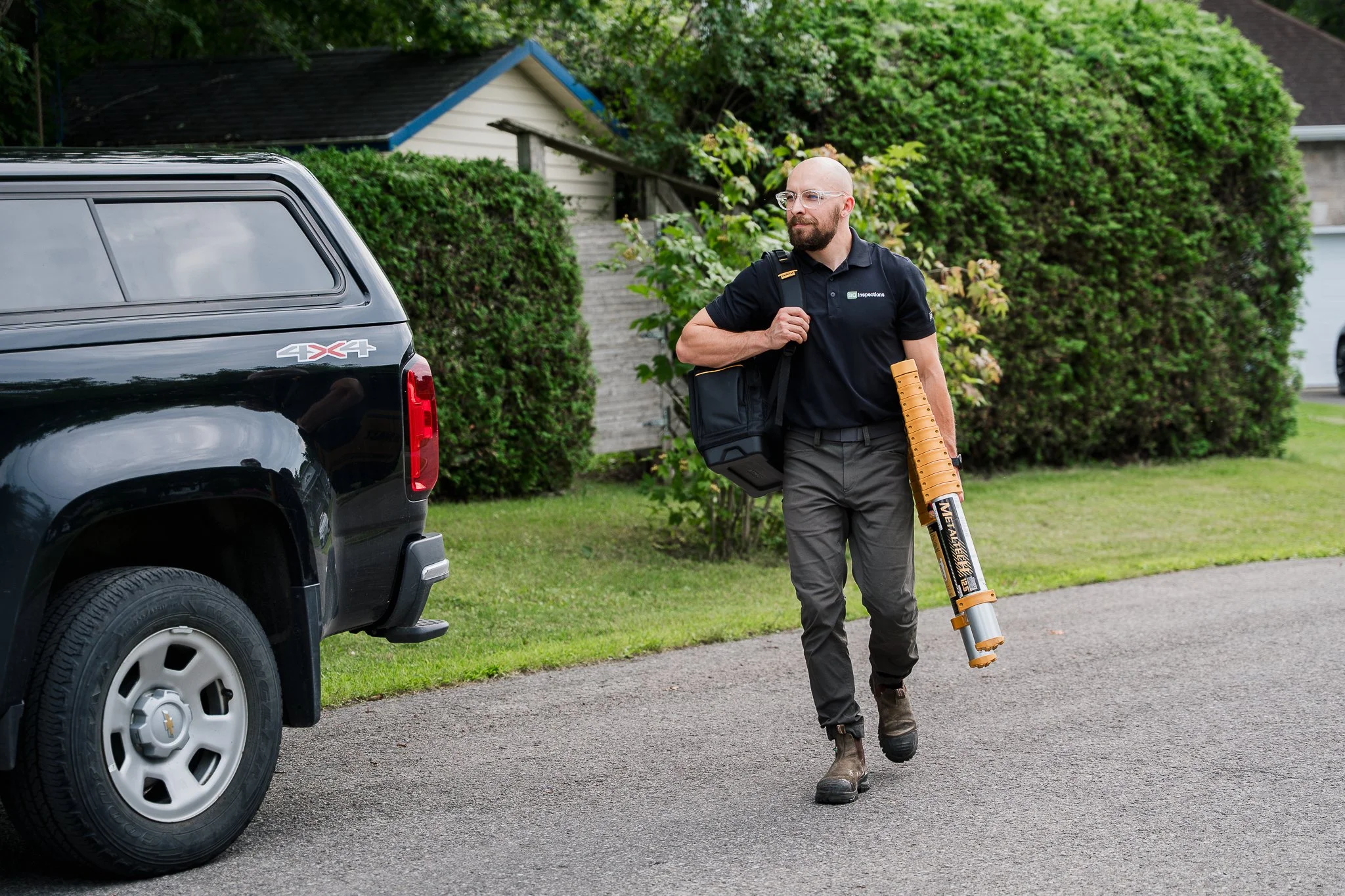 Un homme chauve, portant des lunettes, une chemise noire avec le logo inspecteurs, et un pantalon gris, marche sur le trottoir avec un détecteur de métaux à la main, à côté d'une voiture noire.