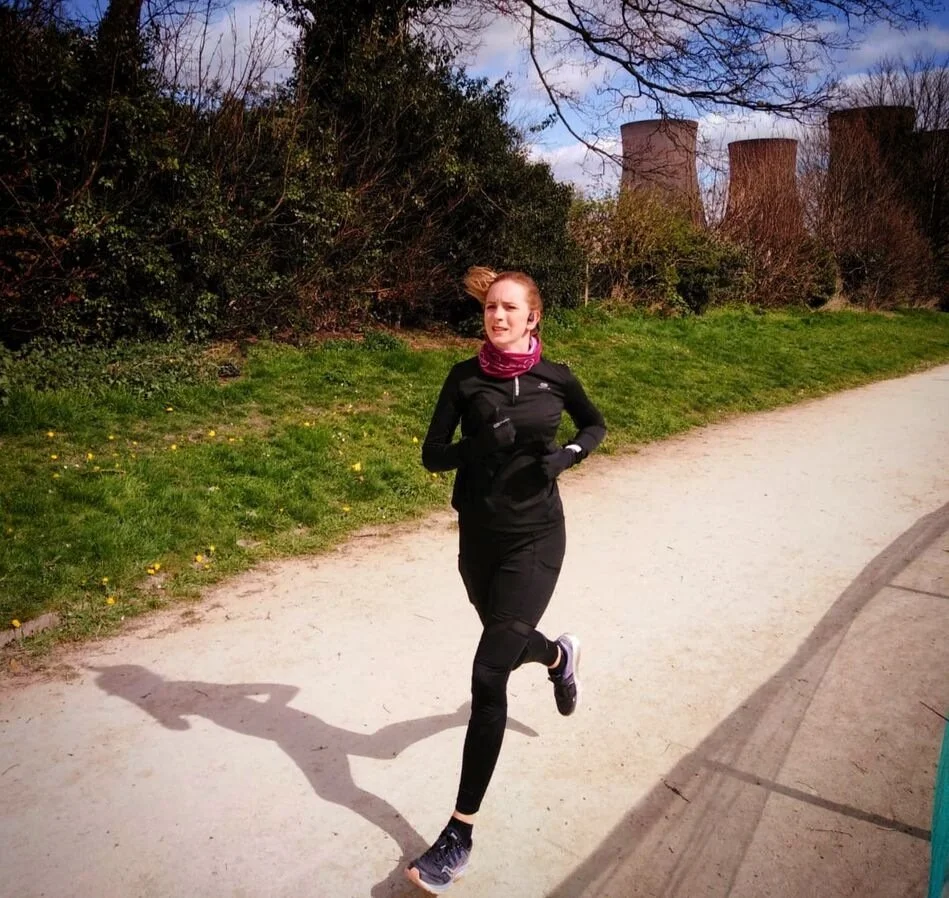 Person jogging on a park path, wearing black athletic clothing, with greenery and cooling towers in the background.