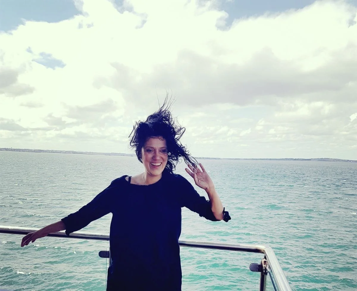 Rani Sheilagh, a woman with curly hair smiling and waving, standing on a boat with an ocean and cloudy sky in the background. Ireland. Cyberpsychologist. Wellness Futurist. Author. Speaker