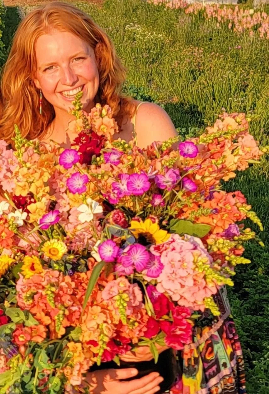 A woman with red hair smiles radiantly with her arms full of wildflowers