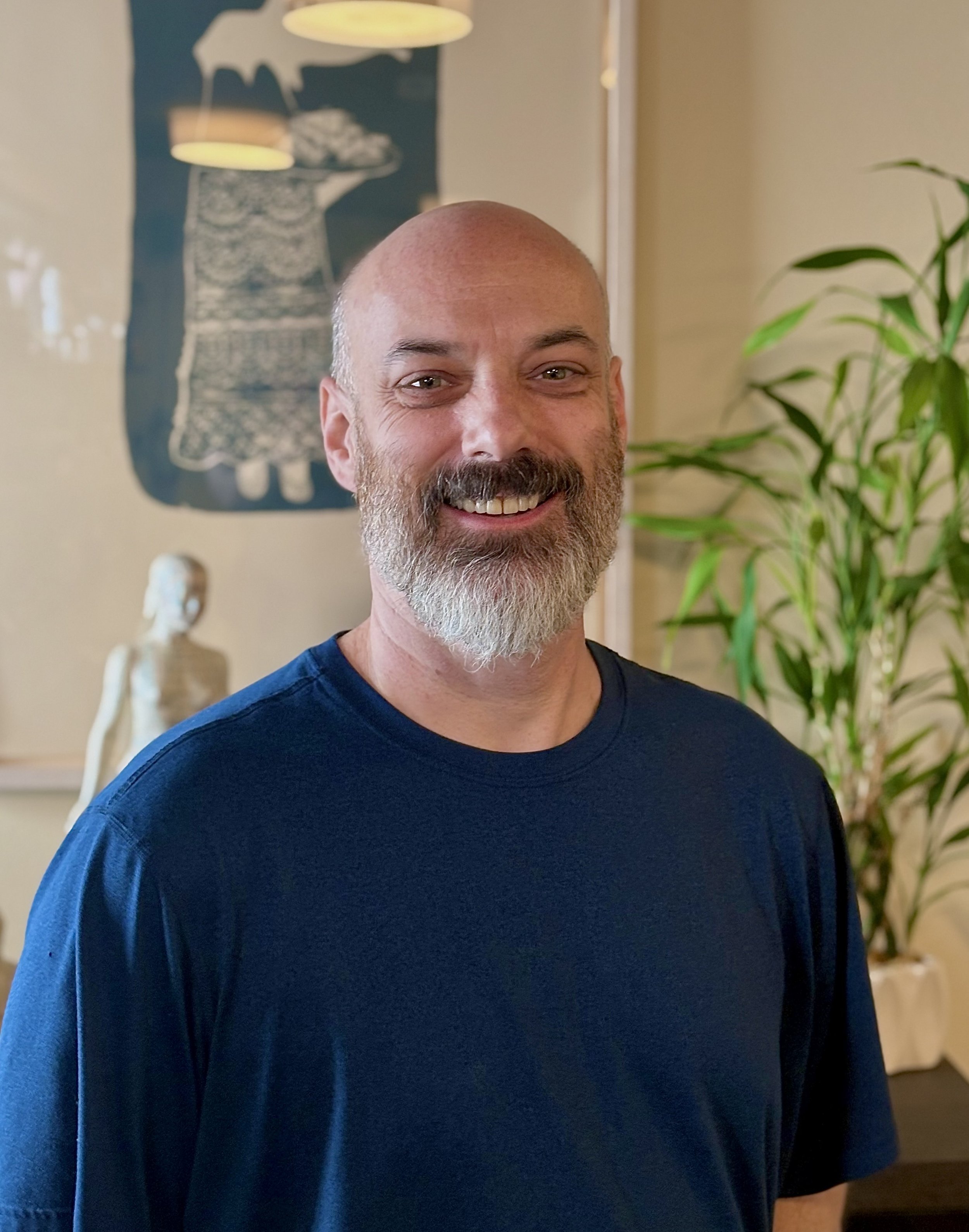 Bald man with a gray beard wearing a blue shirt standing inside of a wellness clinic.
