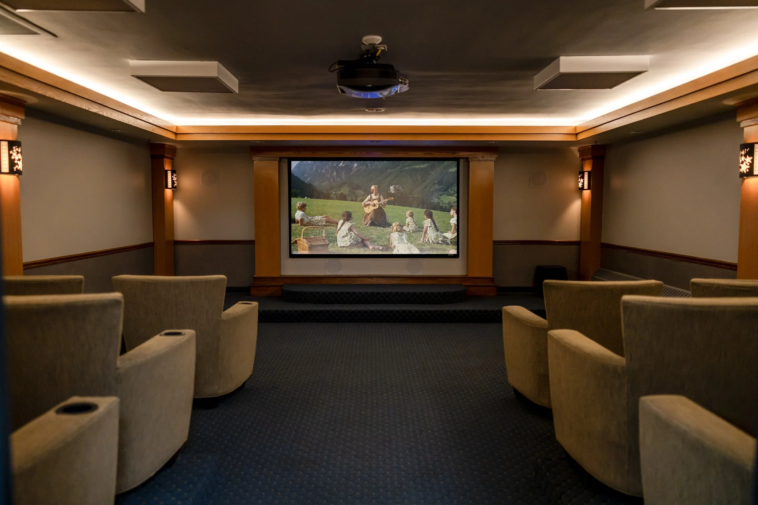 Empty movie theater with beige chairs facing a large screen. The screen displays a scene from "Sound of Music" with characters sitting on grass with mountains in the background, while a person plays guitar.