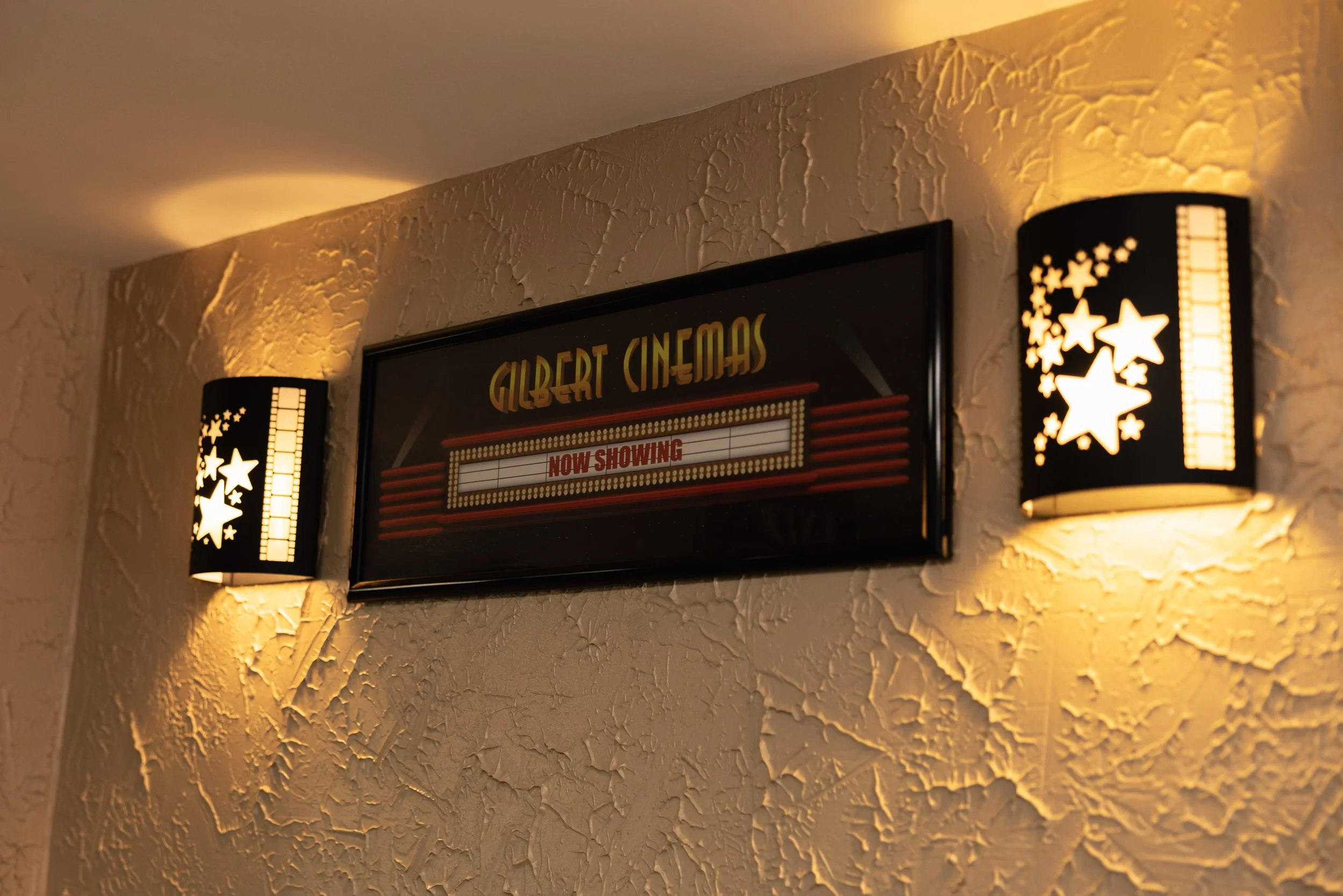 A vintage style cinema sign reading 'Gilbert Cinemas' with a marquee indicating the showtime, flanked by two black wall sconces with star and strip patterns illuminating a textured beige wall.