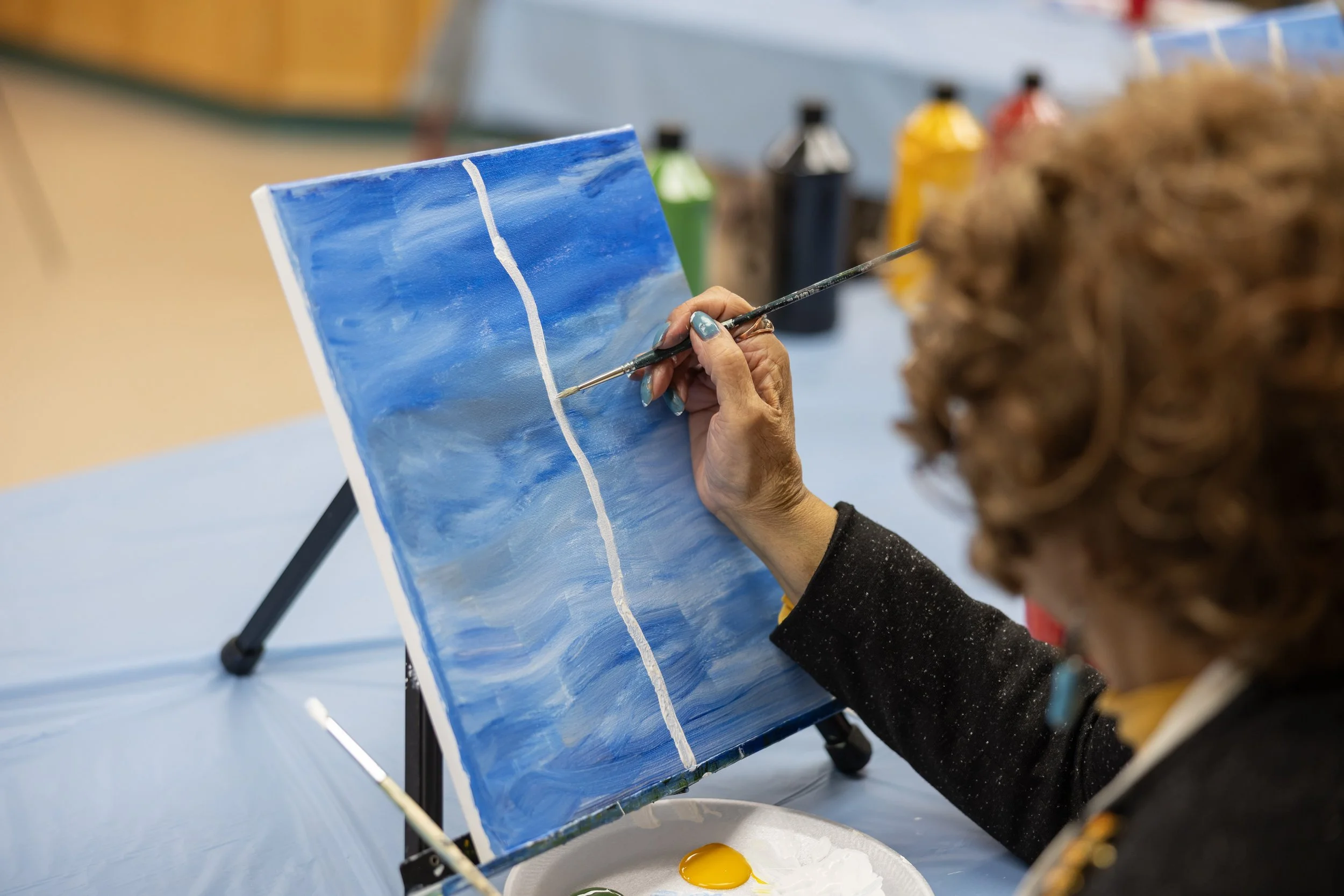 Person painting a landscape on a small canvas with shades of blue, using a thin brush, with bottles of paint in the background.