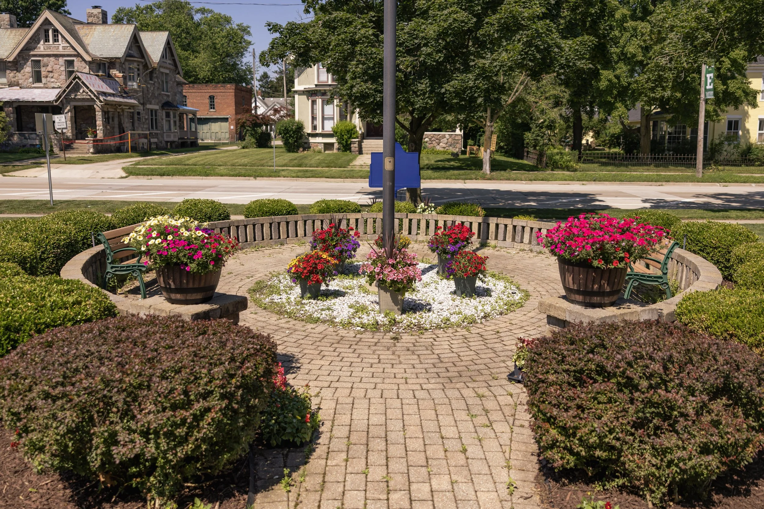 A circular park area with brick pavement, surrounded by green bushes. There are large flower pots filled with colorful flowers, small benches, and a flagpole at the center. Residential houses and trees are visible in the background.