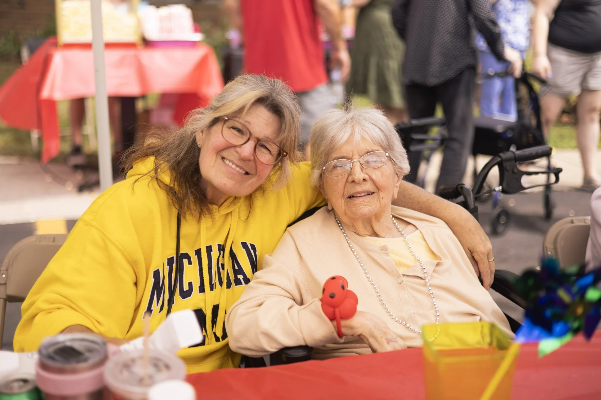 Two women sitting at a table outdoors, one wearing a yellow Michigan hoodie, the other holding a small red balloon, both smiling.