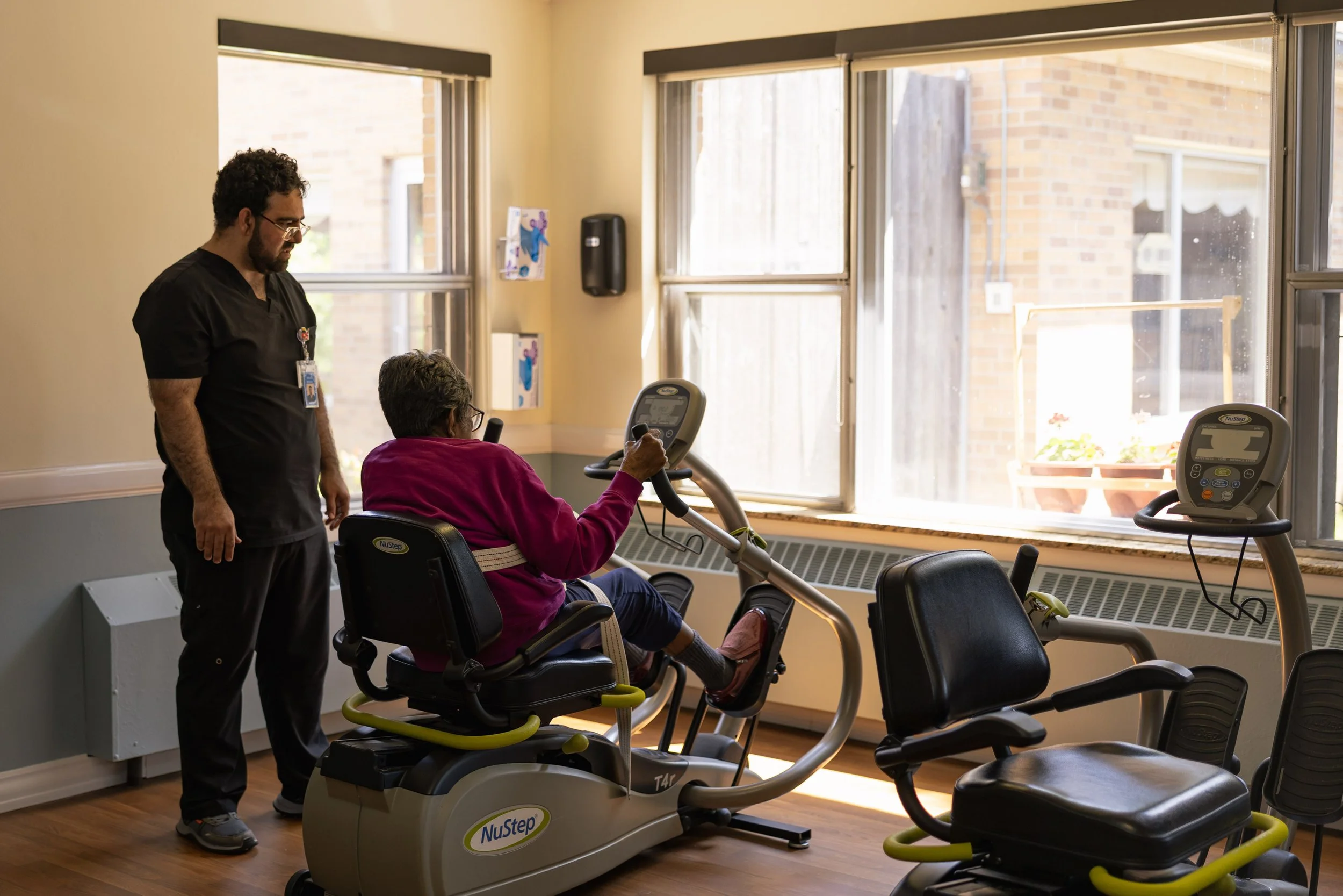 A woman in a pink sweater exercising on a NuStep recumbent cross trainer in a brightly lit room with large windows, accompanied by a Physical Therapist observing her.