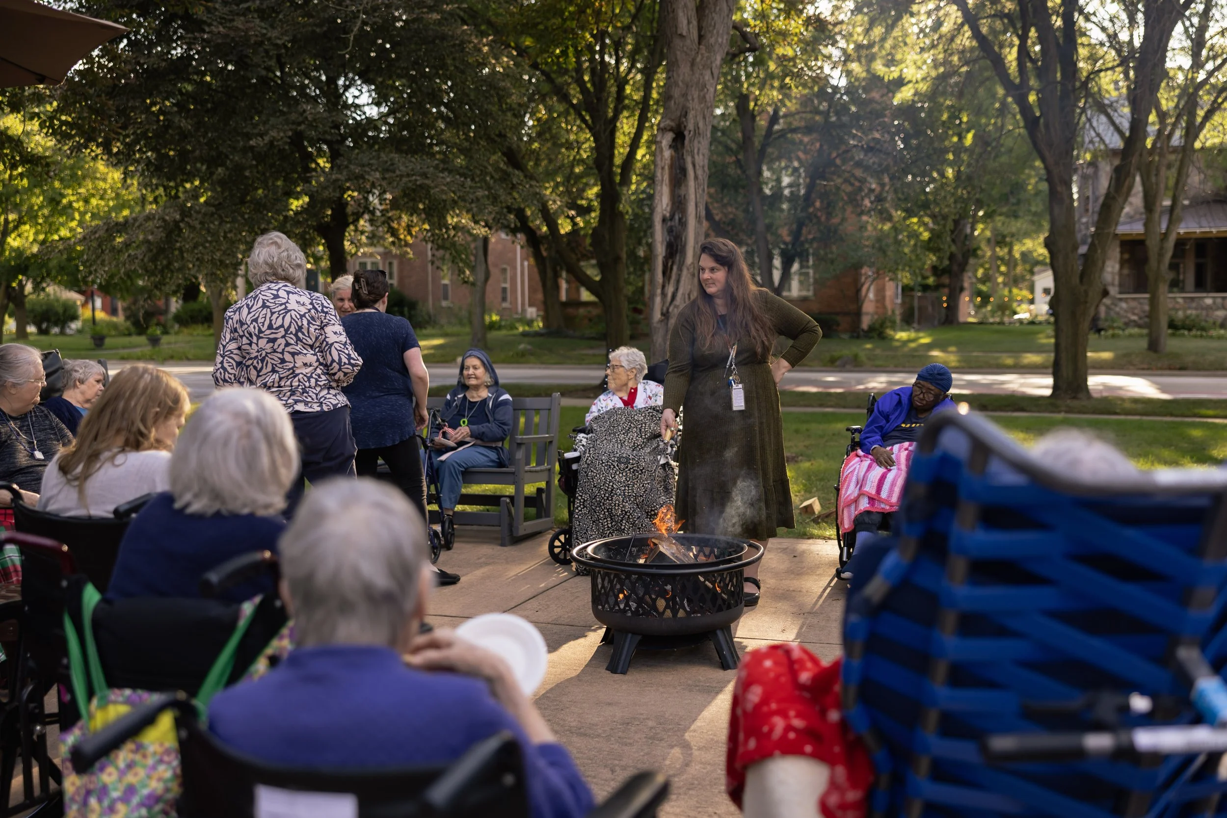 A group of elderly people gathered outside around a small fire pit. Some are sitting in chairs, while others are standing and socializing. The setting appears to be a park or garden with trees and houses in the background during the daytime.