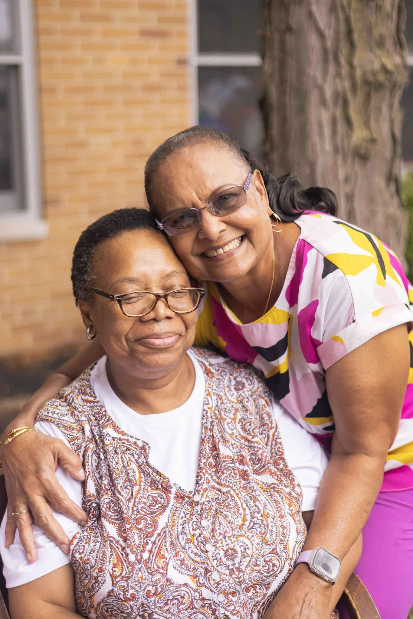 Two women smiling and embracing in front of a brick wall, one wearing a multicolored blouse and glasses, the other wearing a patterned top.