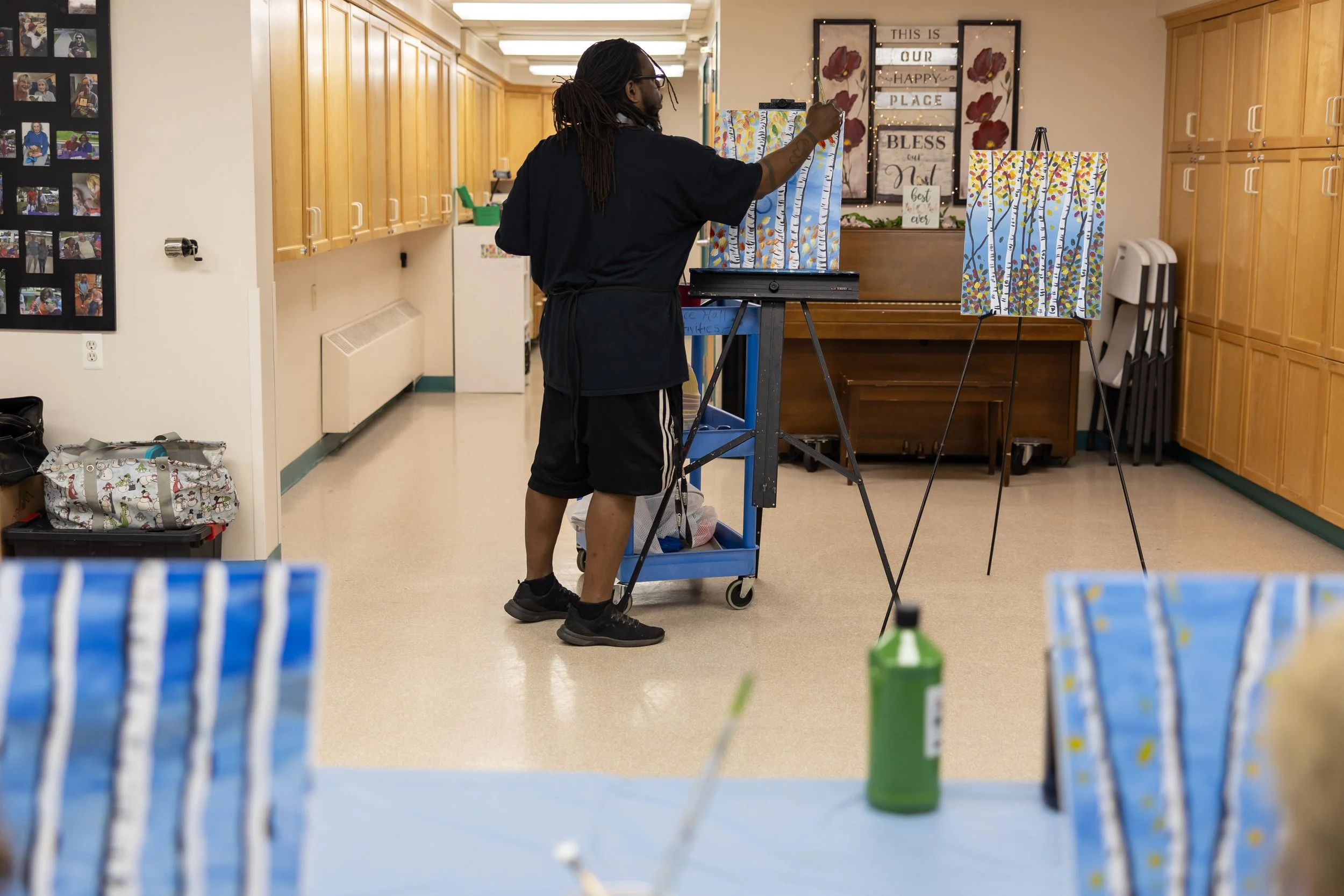 A man painting a colorful landscape on a canvas in an activity room with wooden cabinets and artwork in the background.