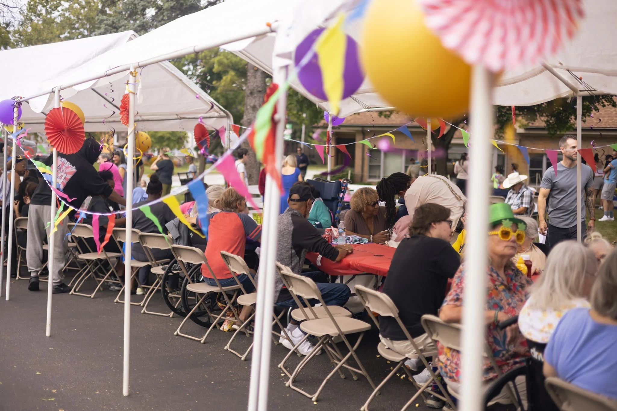 Outdoor community gathering with people seated under tents, decorated with colorful flags and paper ornaments.